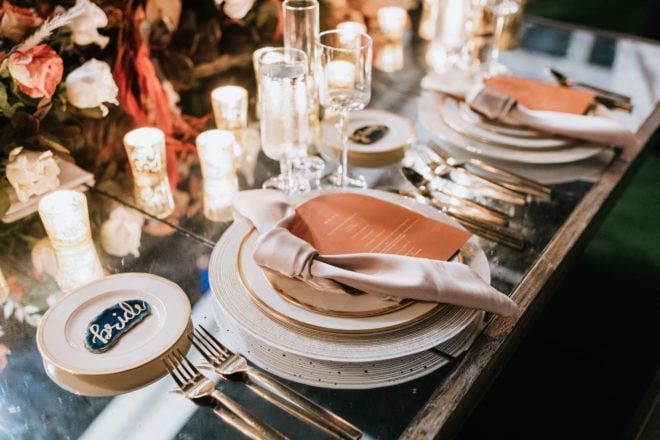 Mirrored tabletop set with white and gold white plates, coral menus with rounded edges, agate stones with the words "bride" and "groom" written on them in gold calligraphy and lit gold votives amidst white, coral and blush colored florals provided by Plants N Petals.