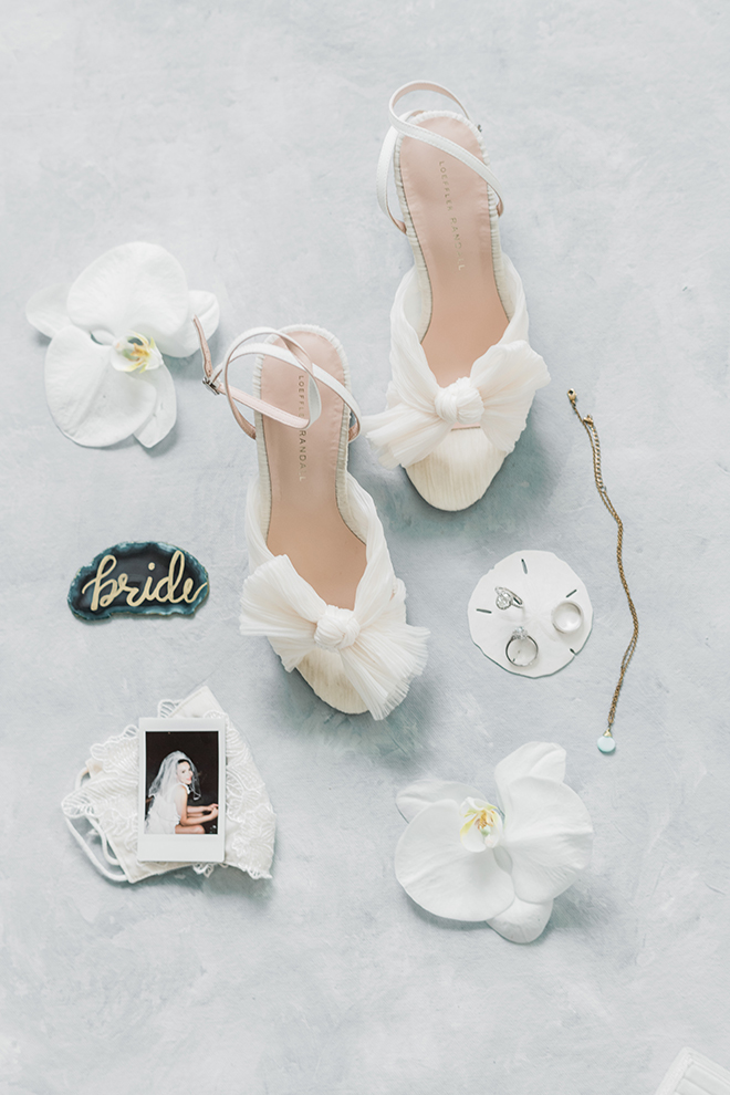 Loeffler Randall knotted bow sandals sit next to a polaroid of the brides mother, a teal slice of agate, and the wedding bands resting on a sand dollar.