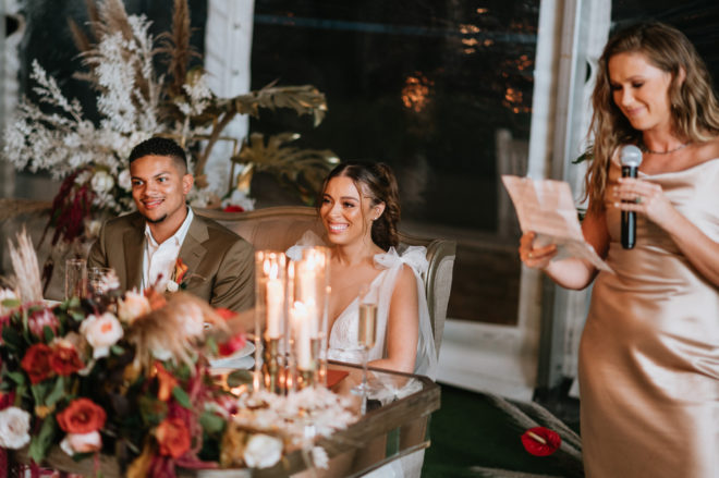 Smiling Groom and bride sitting at a mirrored head table next to the maid of honor wearing a silk blush dress holding a microphone and a piece of paper during her reception speech.