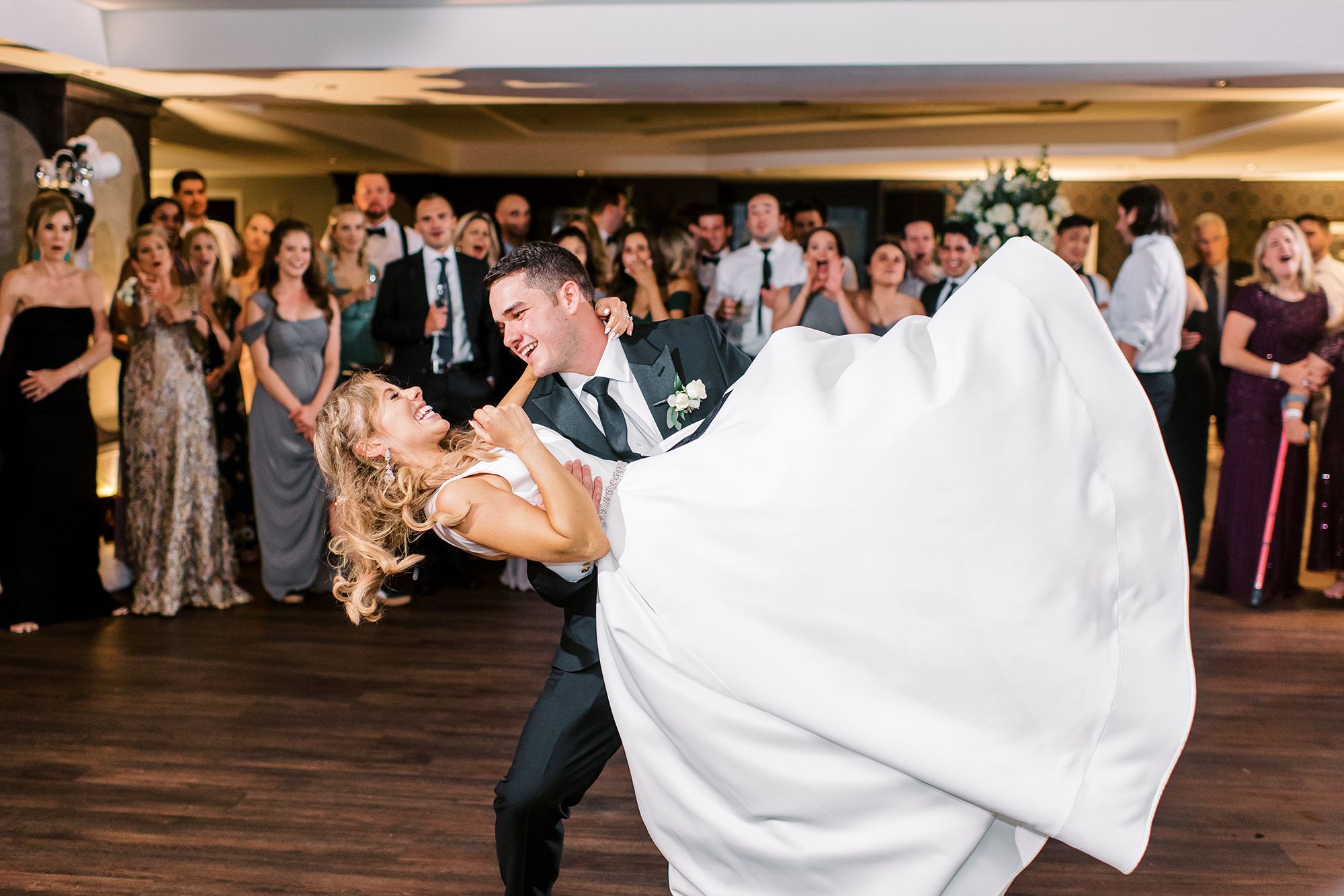 The groom dips the bride on the dance floor while their guests circle them and cheer them on.