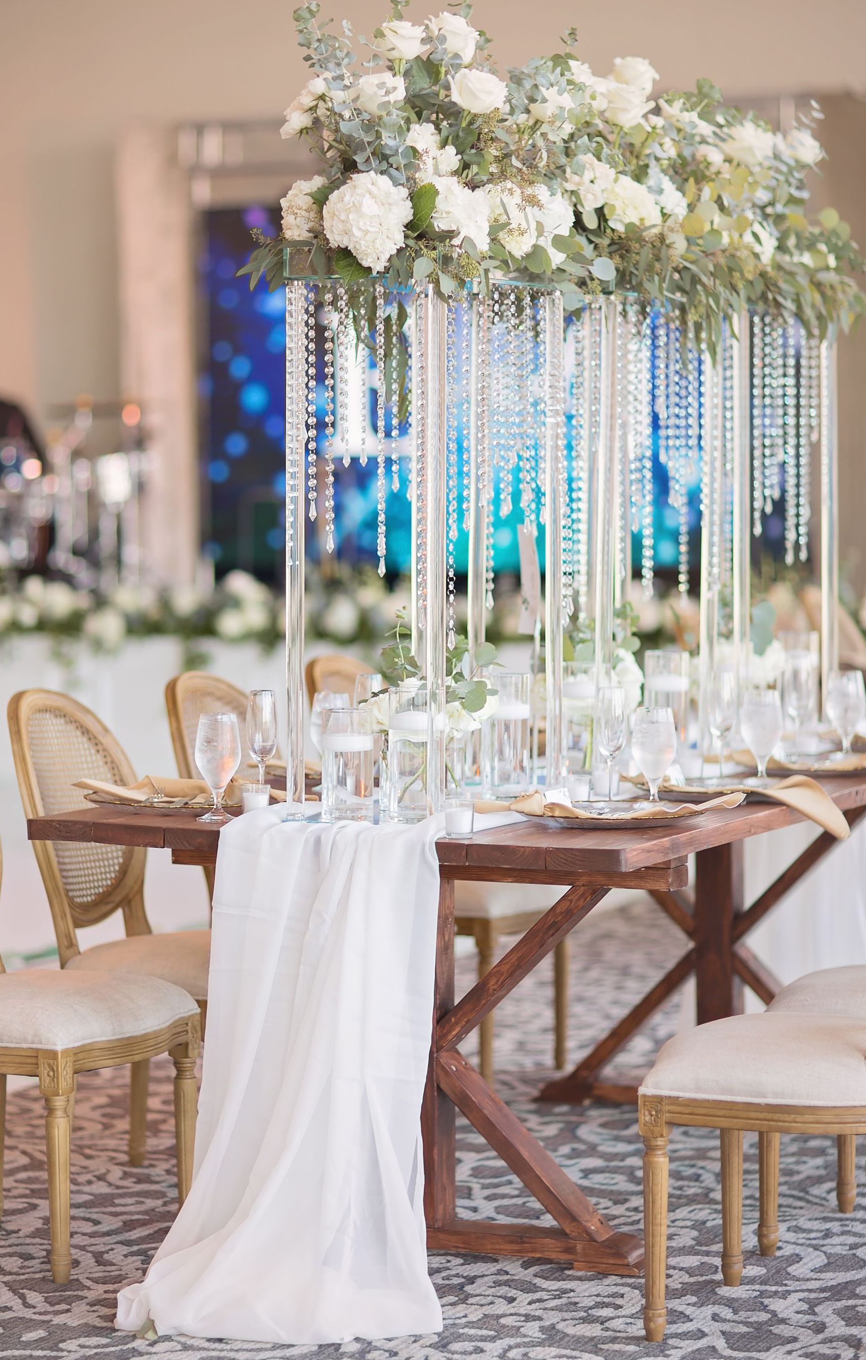 Crystal glass reception decor is set up in the center of the table, including a long white table runner, floating candles, small singular candles, white rose and eucalyptus floral arrangements, and hanging crystal beads. 