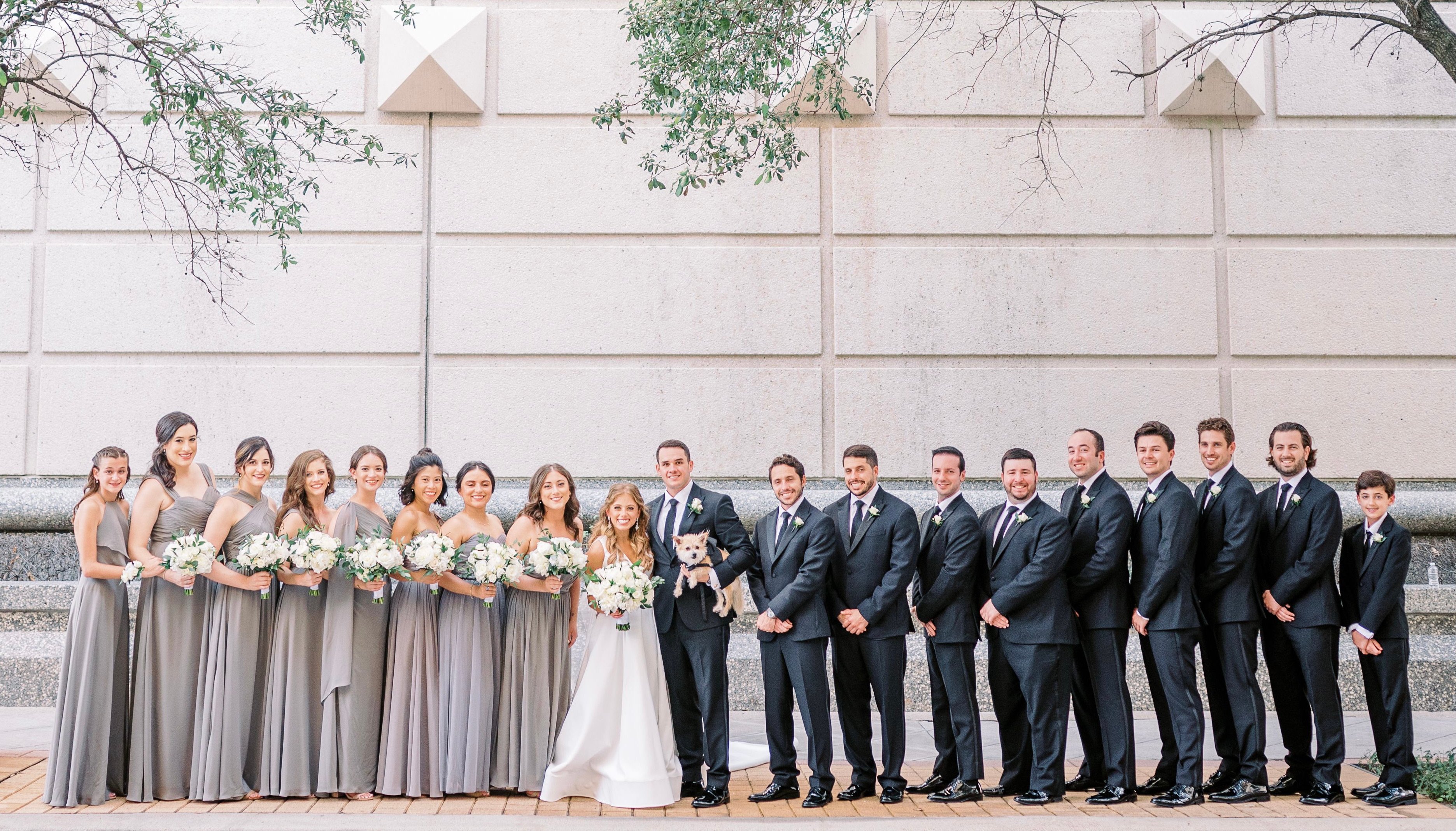 Bride and groom stand outside the venue in Downtown Houston with all of their bridesmaids and groomsmen. The groom is also holding his dog. The wedding party all wear monochromatic colors of white, gray and black.