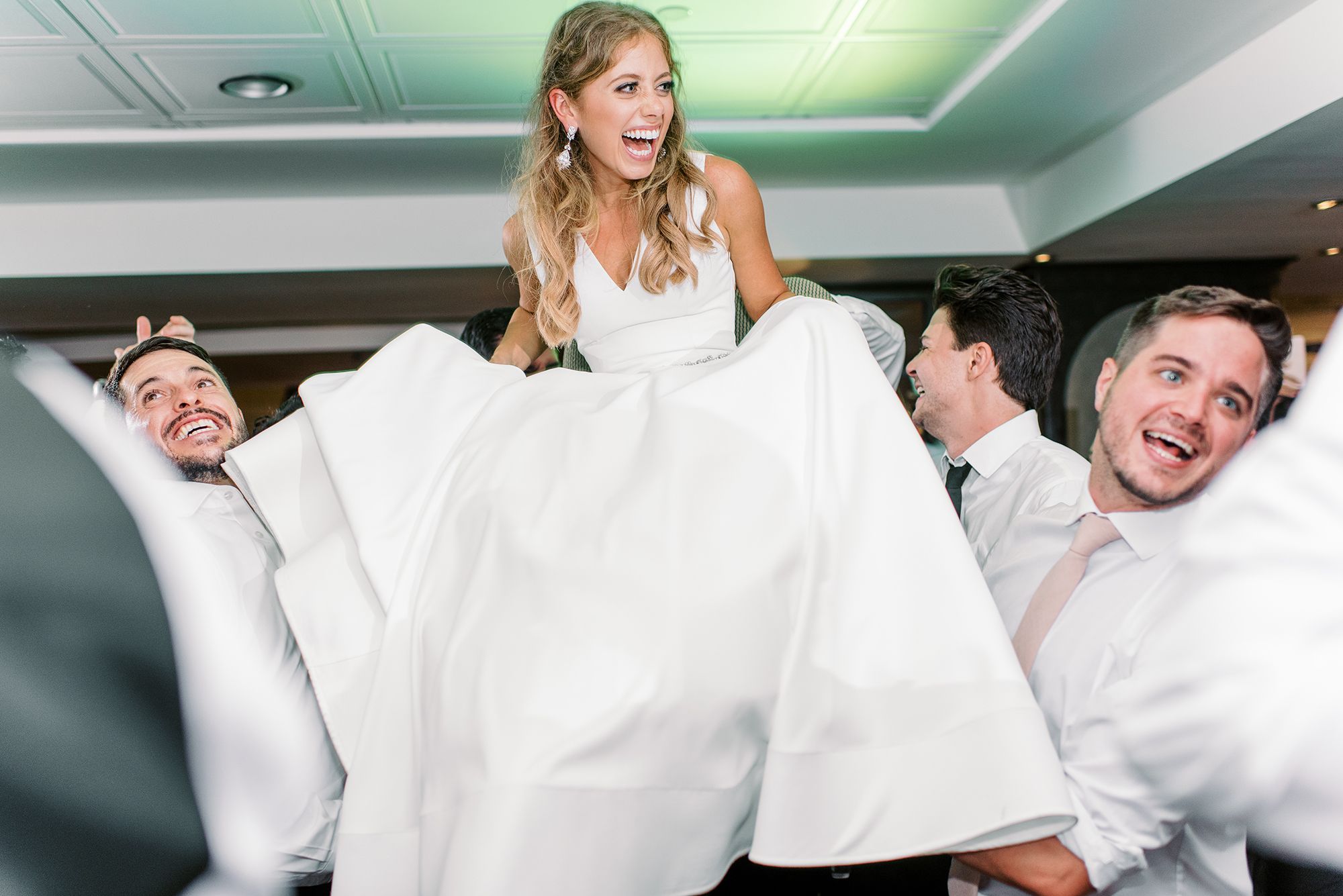 The bride smiling as her wedding guests lift her in a chair at the reception party.
