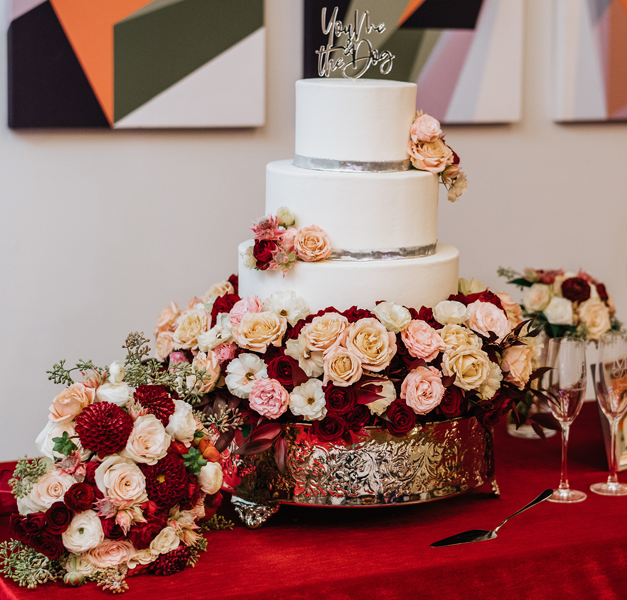 The wedding cake is surrounded with ruby red roses and beige and pink filler flowers.