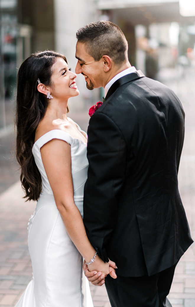 The bride and groom hold hands and smile at each other outside of JW Marriott Downtown Houston. The groom has a ruby red boutonniere. 