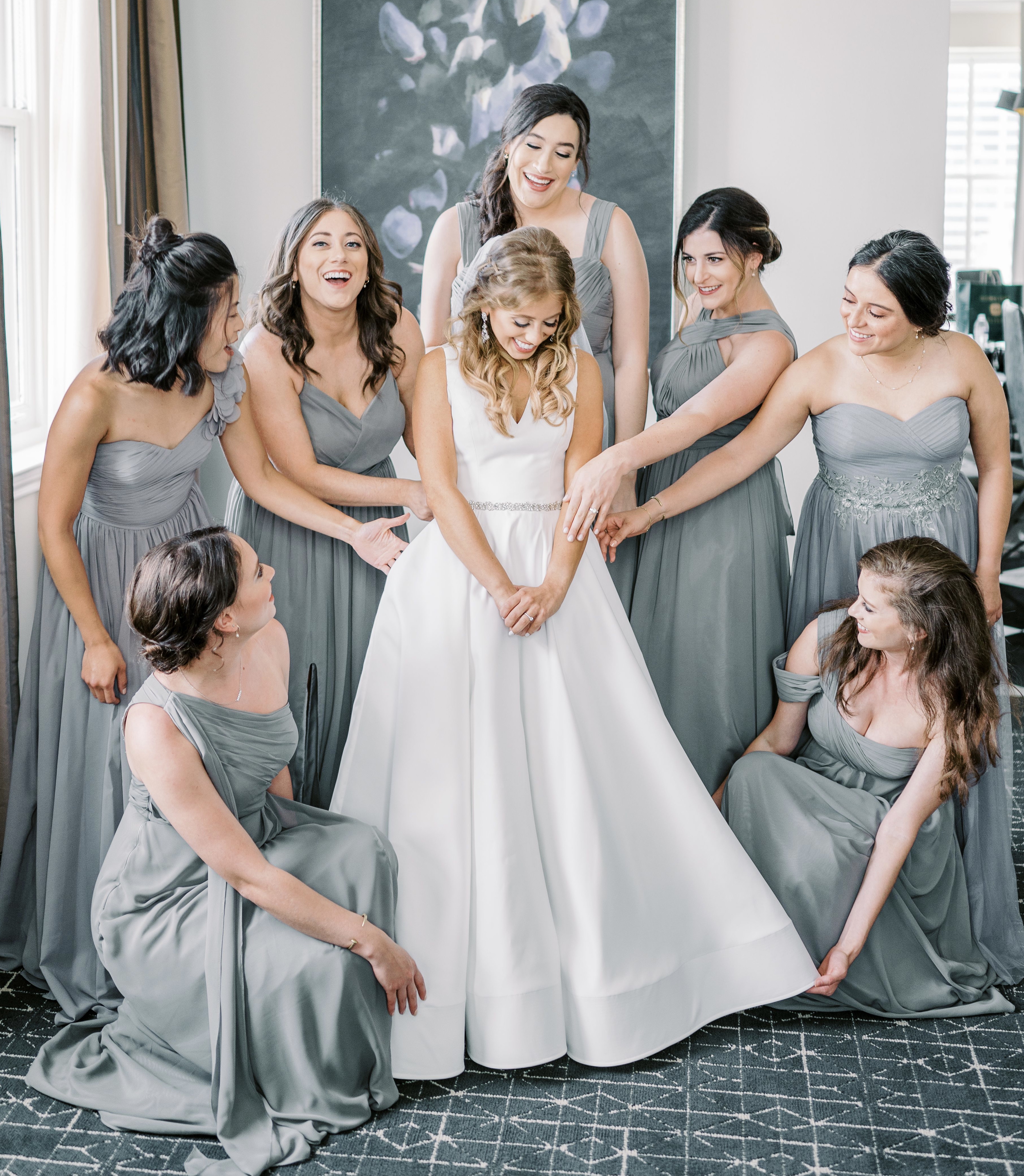 The bride is in her white wedding gown smiling and looking down at the floor while her bridesmaids surround her in monochromatic gray dresses and admire her.