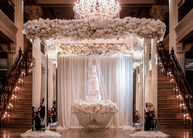 A six tiered cake is on a floral decorated table in the center of a pillared white floral decor set up and illuminated by the chandelier above it. 