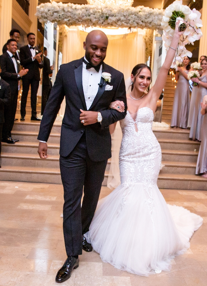 Bride lifts her bouquet up in one hand and she and her groom embrace and celebrate while they walk down the aisle. 