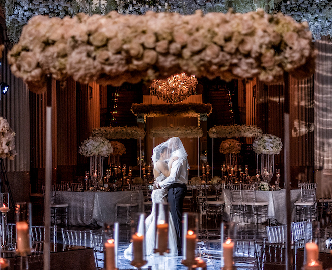 Bride dances with a groom with her long veil over both their heads in a dimly lit Grand Hall decorated with candlelights, white roses and tall centerpieces at the downtown Houston wedding venue, Corinthian Houston.