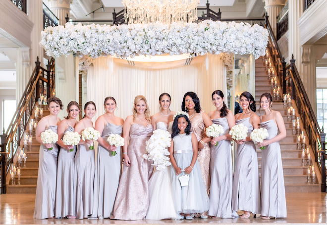 Bride and bridesmaids under a canopy of white lush florals in front of a grand staircase at The Corinthian Houston. 