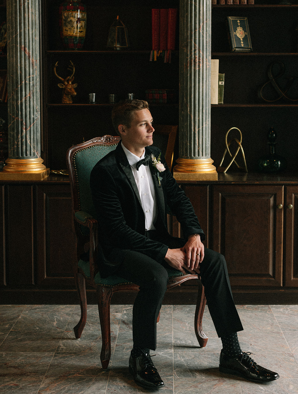 Groom looks out the window of the venue The Mansion at ColoVista while sitting in a green chair.