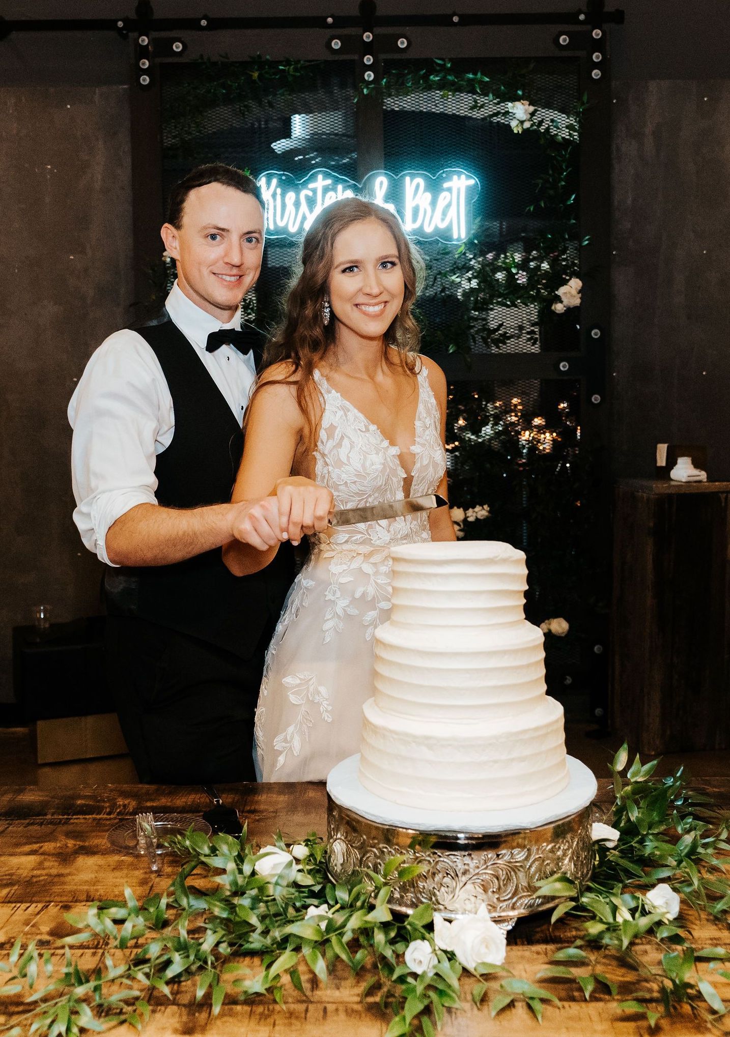 Kirsten and Brett prepare to cut their three-tiered plain white cake while a neon sign with the couple's names shines bright in the background.