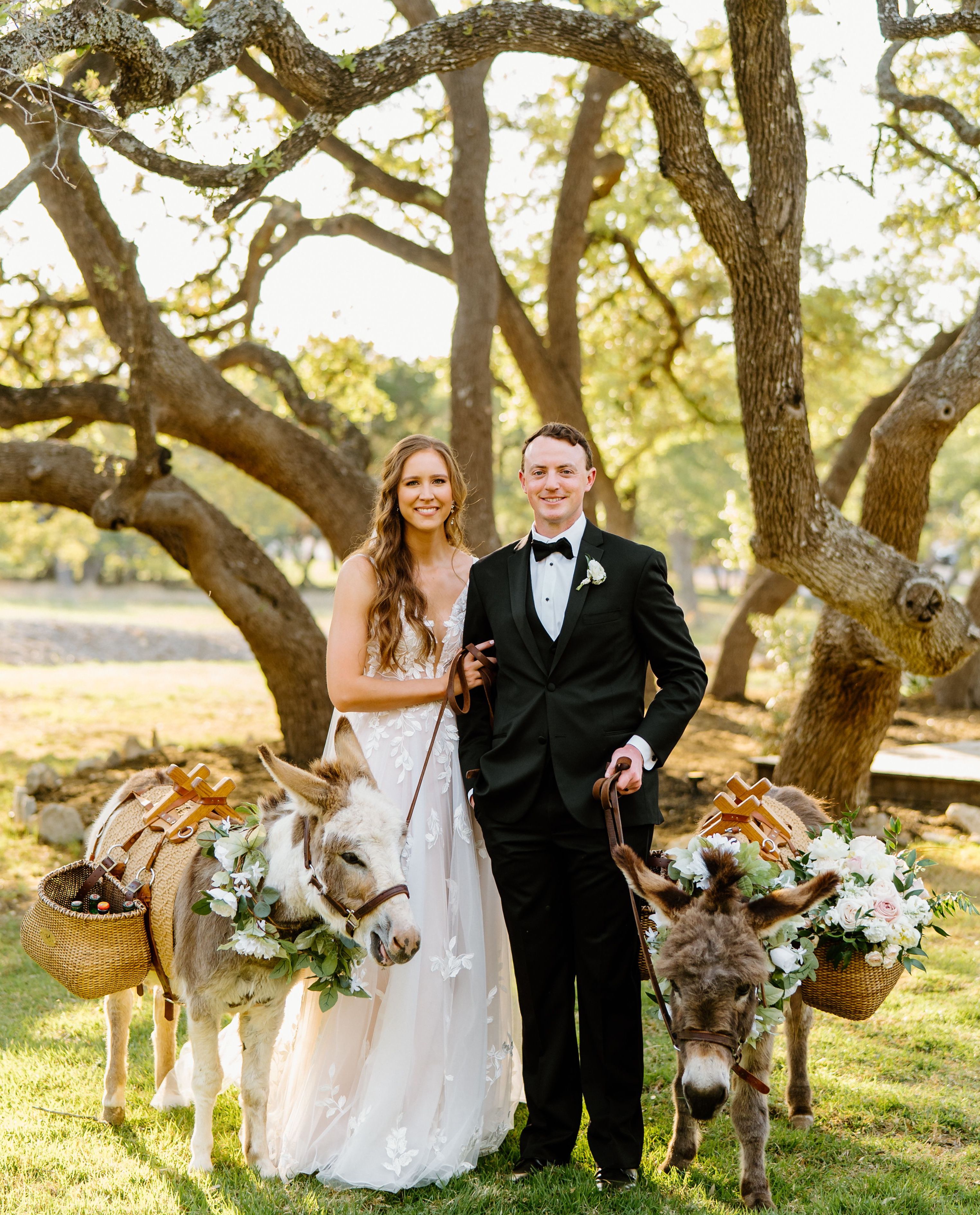 A smiling bride and groom are flanked by two floral adorned beer burros with a towering oak tree behind them.