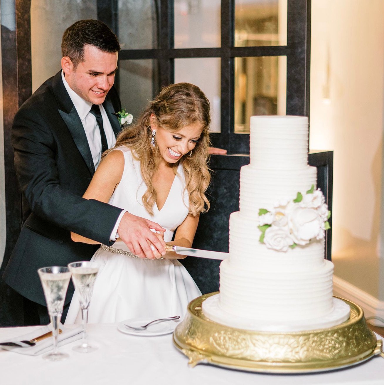 Bride and groom cut into their classic white, 4-tiered wedding cake while smiling.