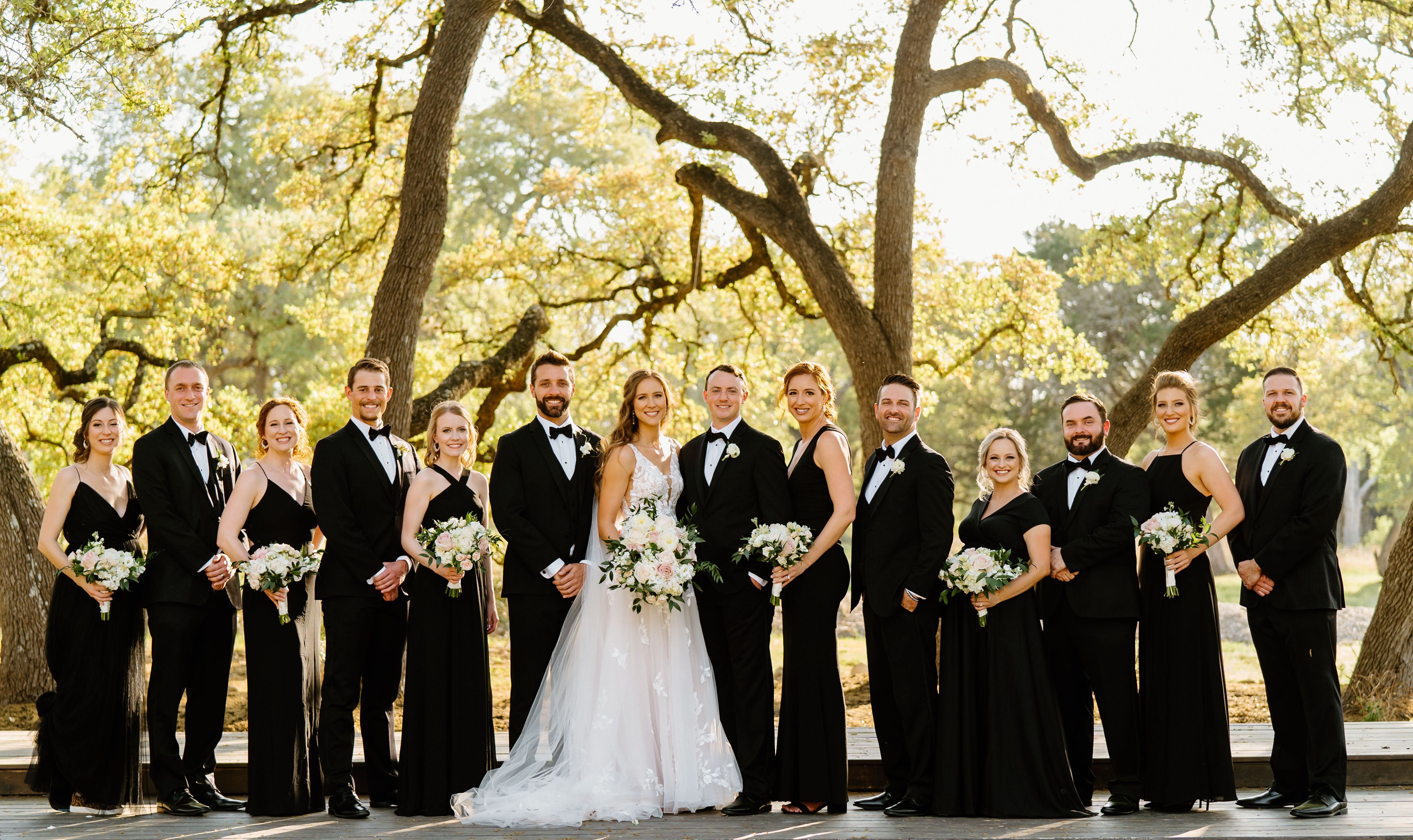 A bride and groom stand in the center of their wedding party adorned in black tie attire after the wedding ceremony with trees and natural light in the background.