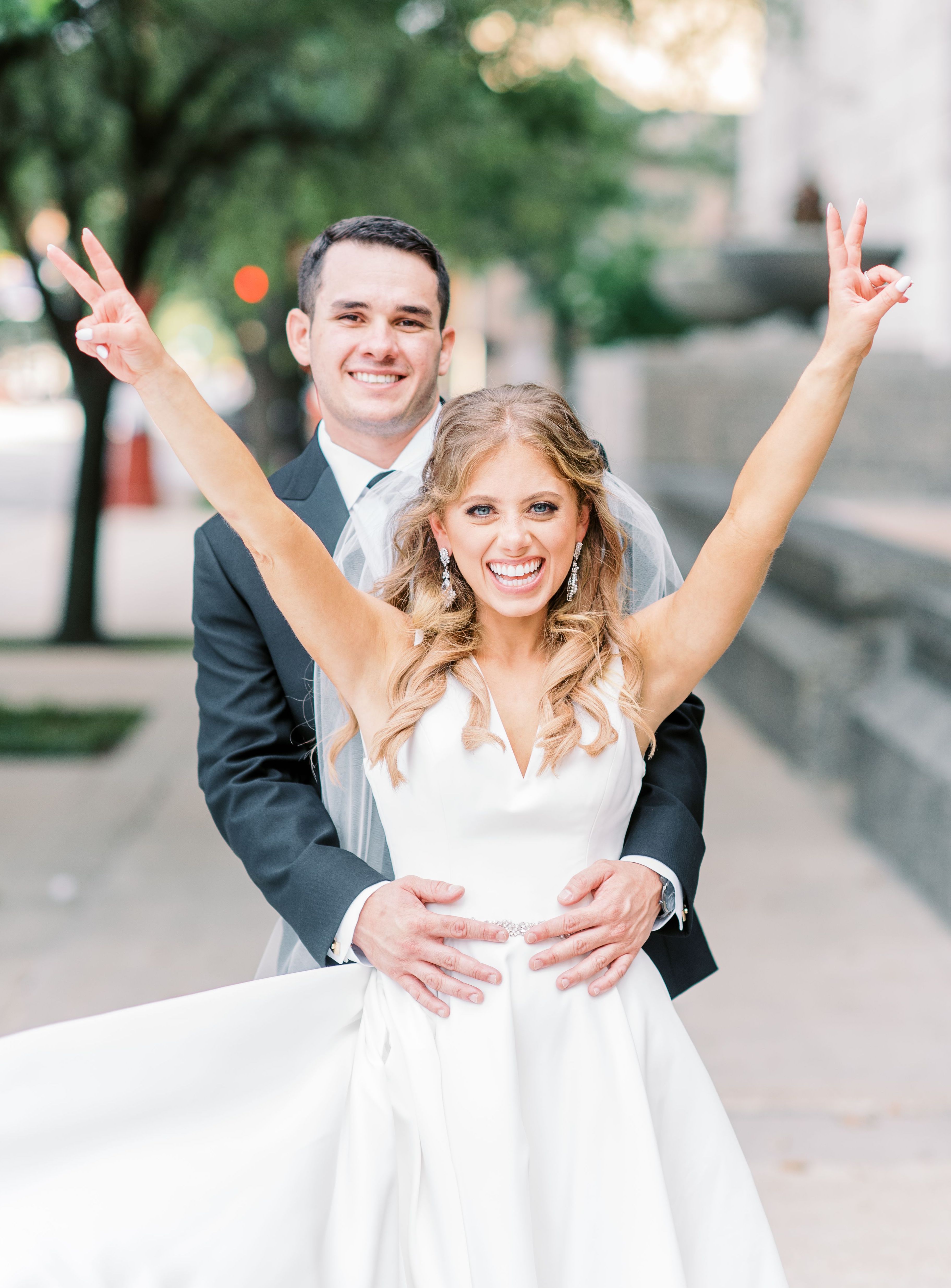 Bride holding up peace signs with her hands and smiling as the groom holds her waist.