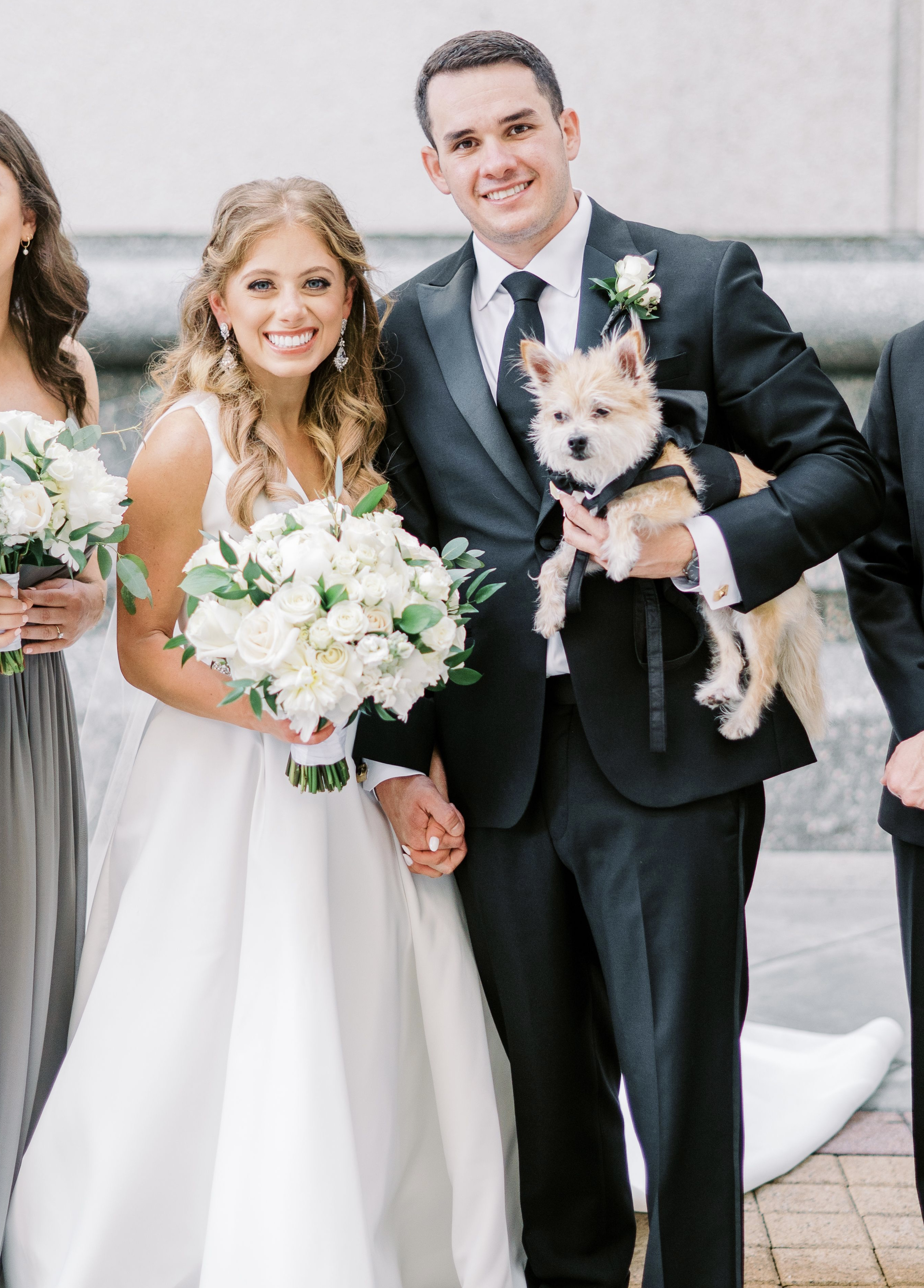 Bride and groom are holding hands while the groom holds their dog who is wearing a black tux.