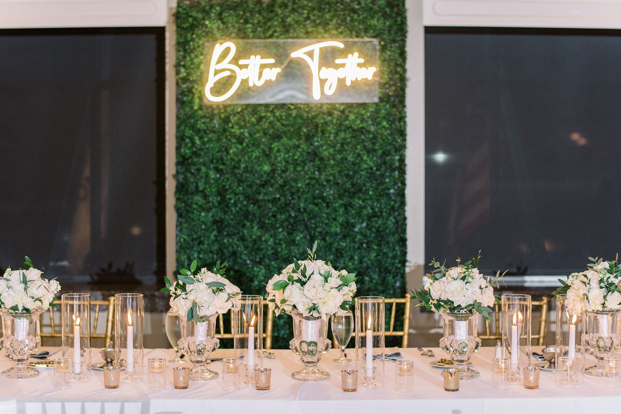 The bride and groom's table is decorated with floral centerpieces with white flowers. The backdrop is an ivy wall with a neon sign that says "Better Together."