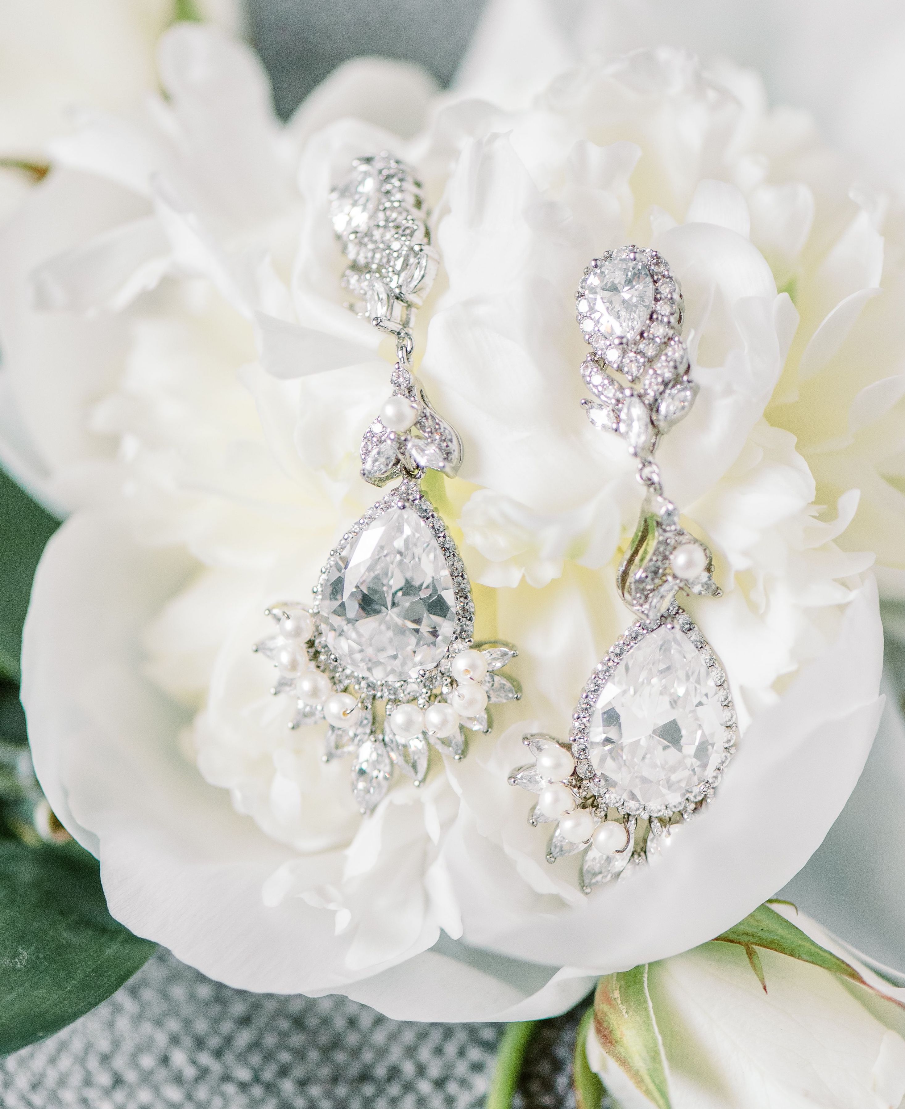 Sparkling diamond dangle earrings with classic small pearls embellished as well laying on a white peony.