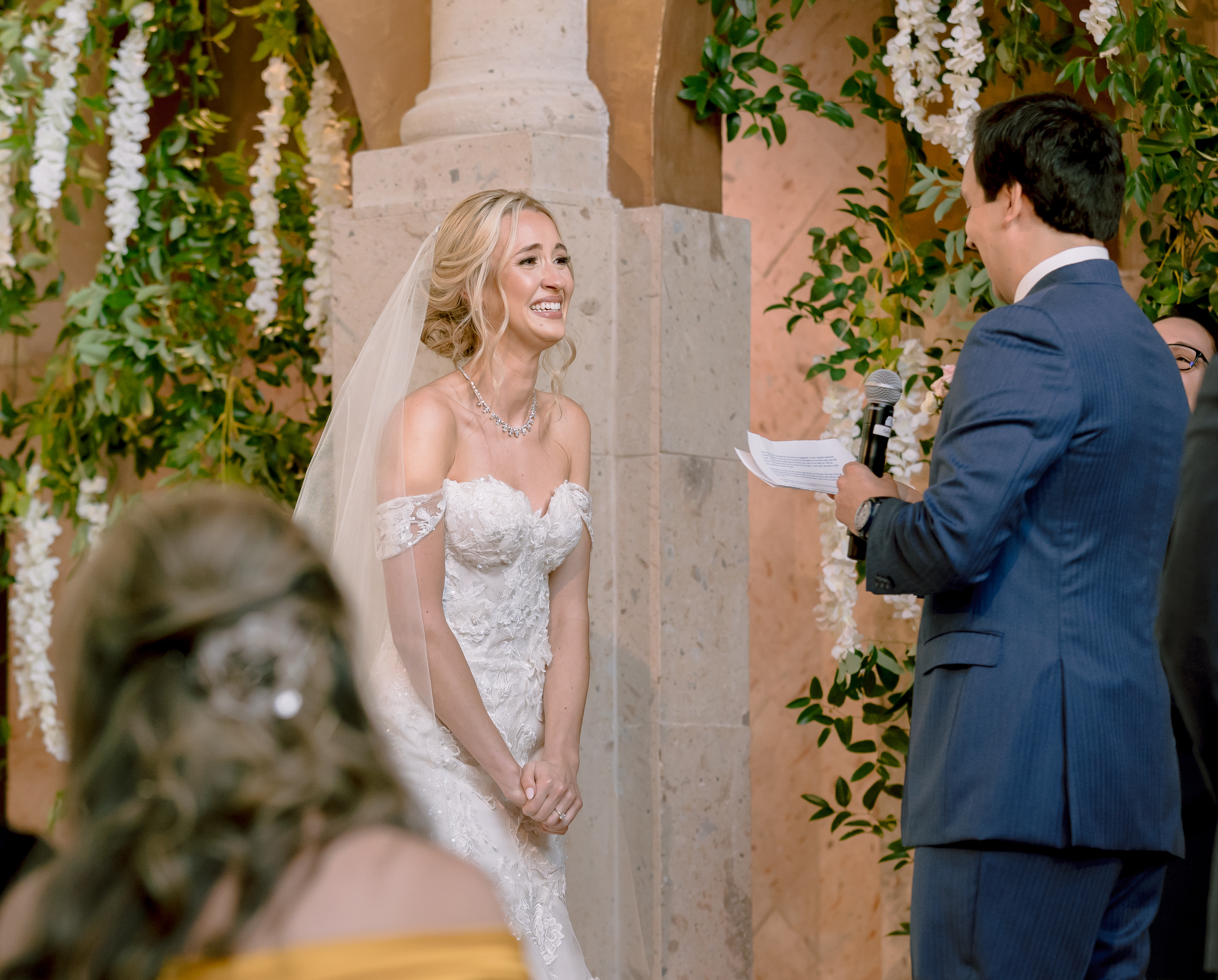 The bride smiles at her groom as he recites his vows at their greenery-filled wedding.