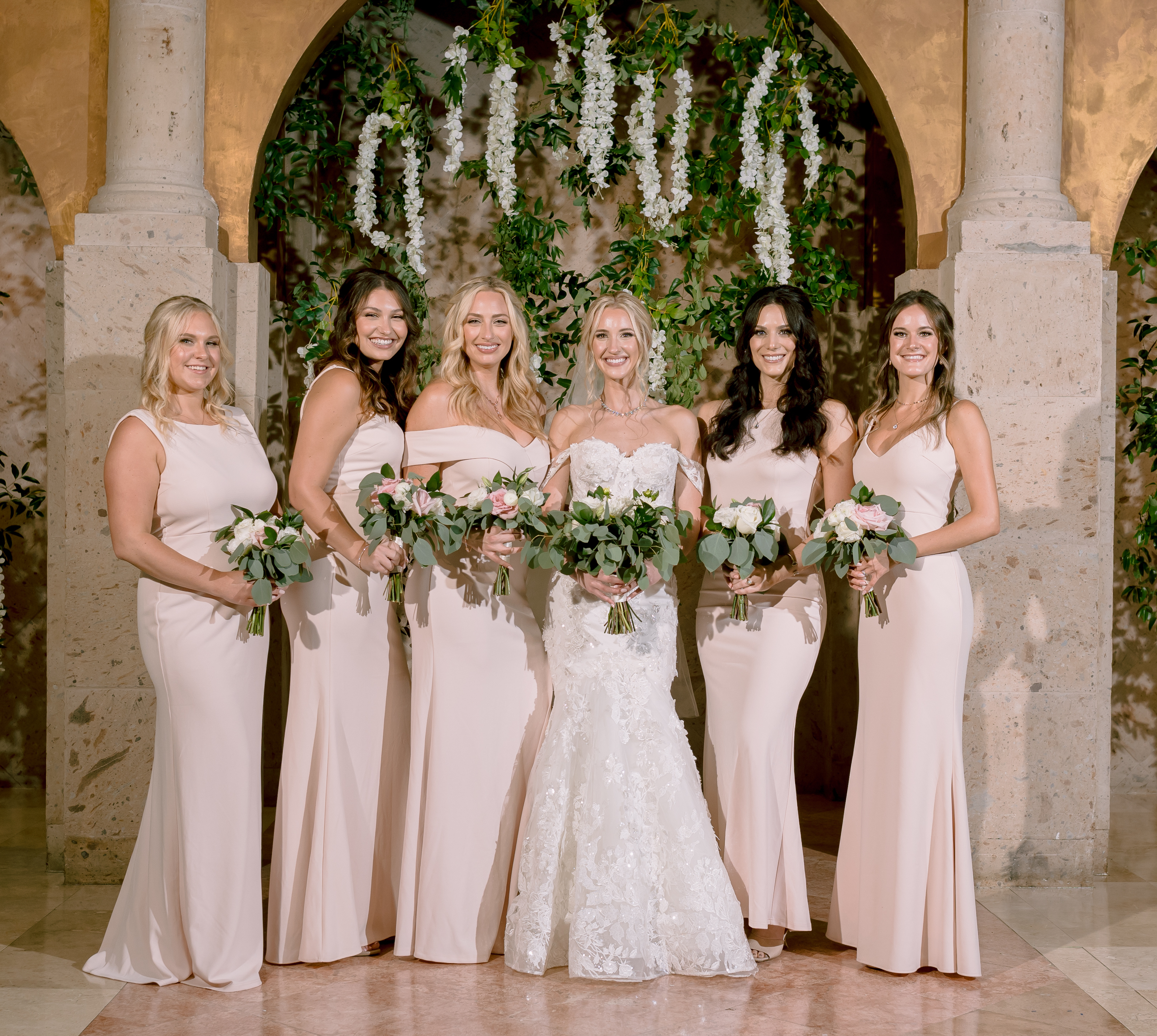 The bride and her bridesmaids smile in front of the wisteria at her greenery-filled wedding.
