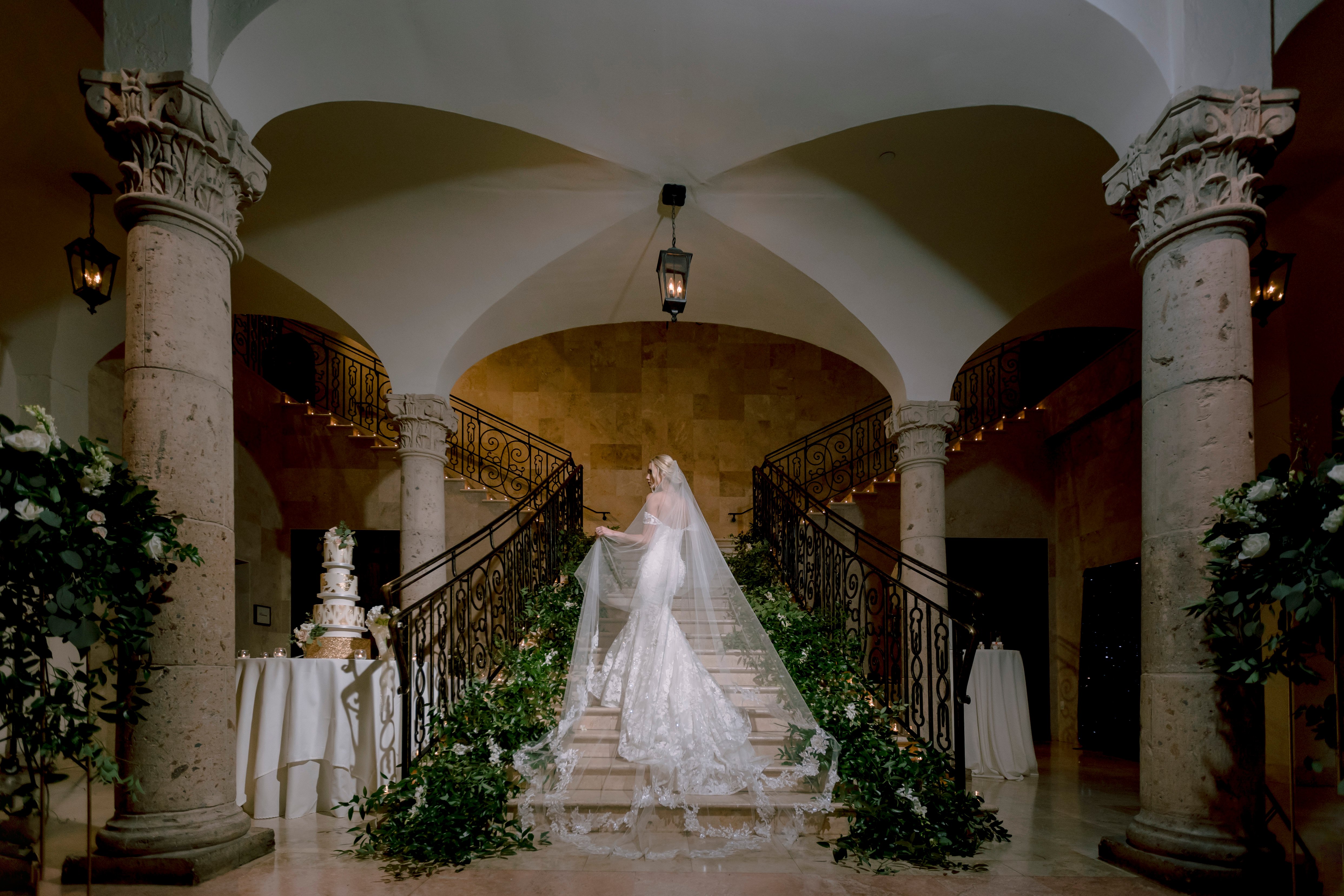 The bride poses in the middle of the grand staircase with her veil trailing behind her before her greenery-filled wedding.