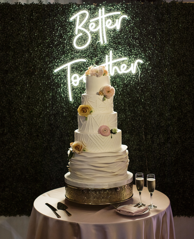 The wedding cake with a neon sign behind it that says " Better Together."