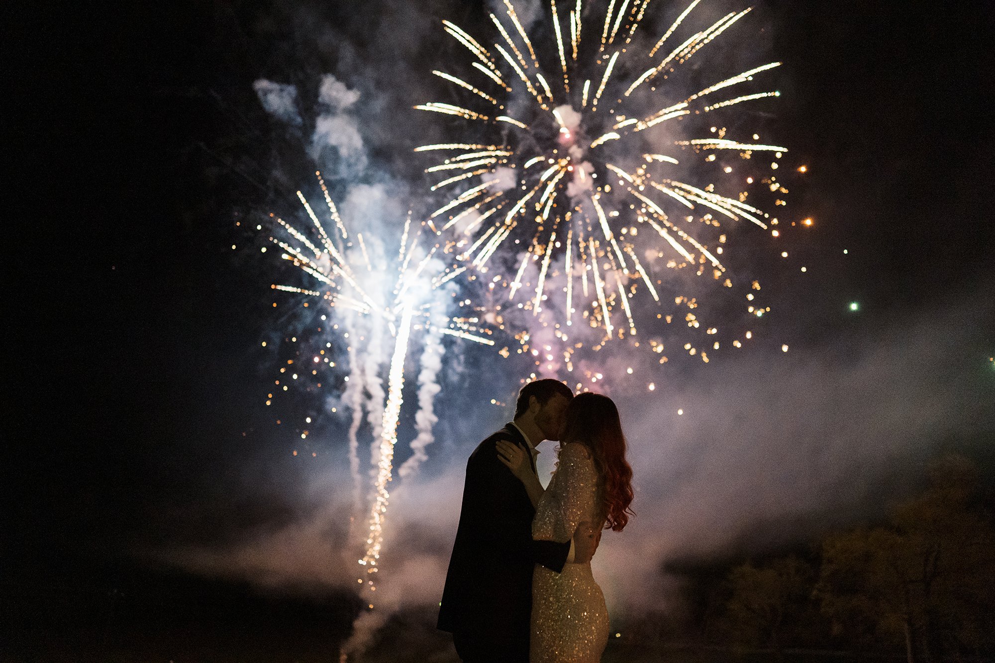 The bride and groom kiss with fireworks exploding behind them.