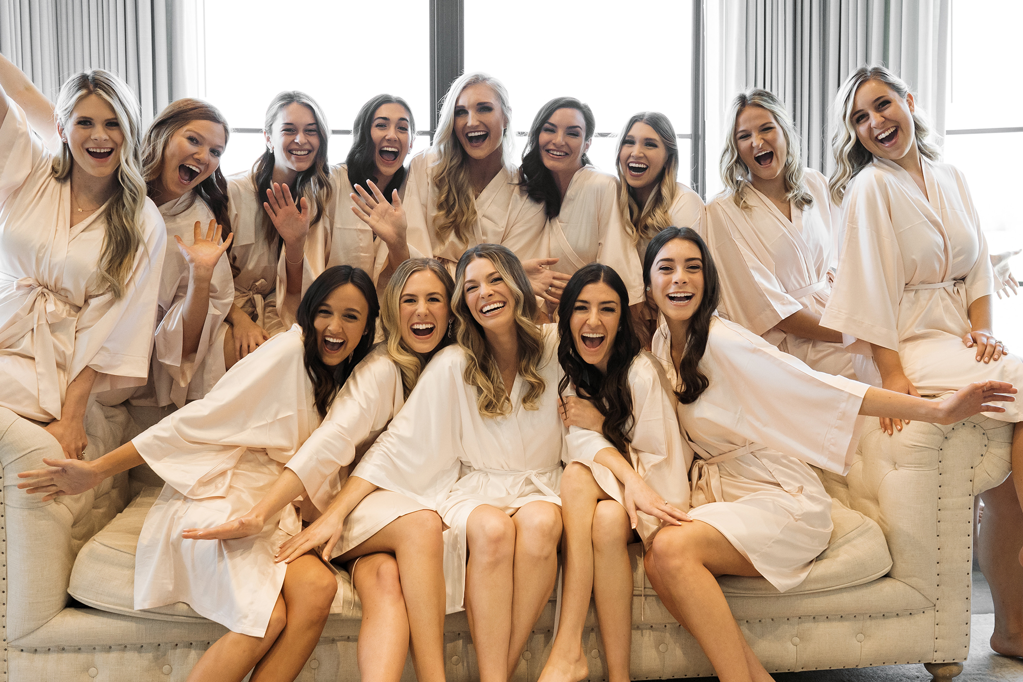 The bride and her bridesmaids pose and smile while sitting on a couch in the bridal suite for their timeless wedding.