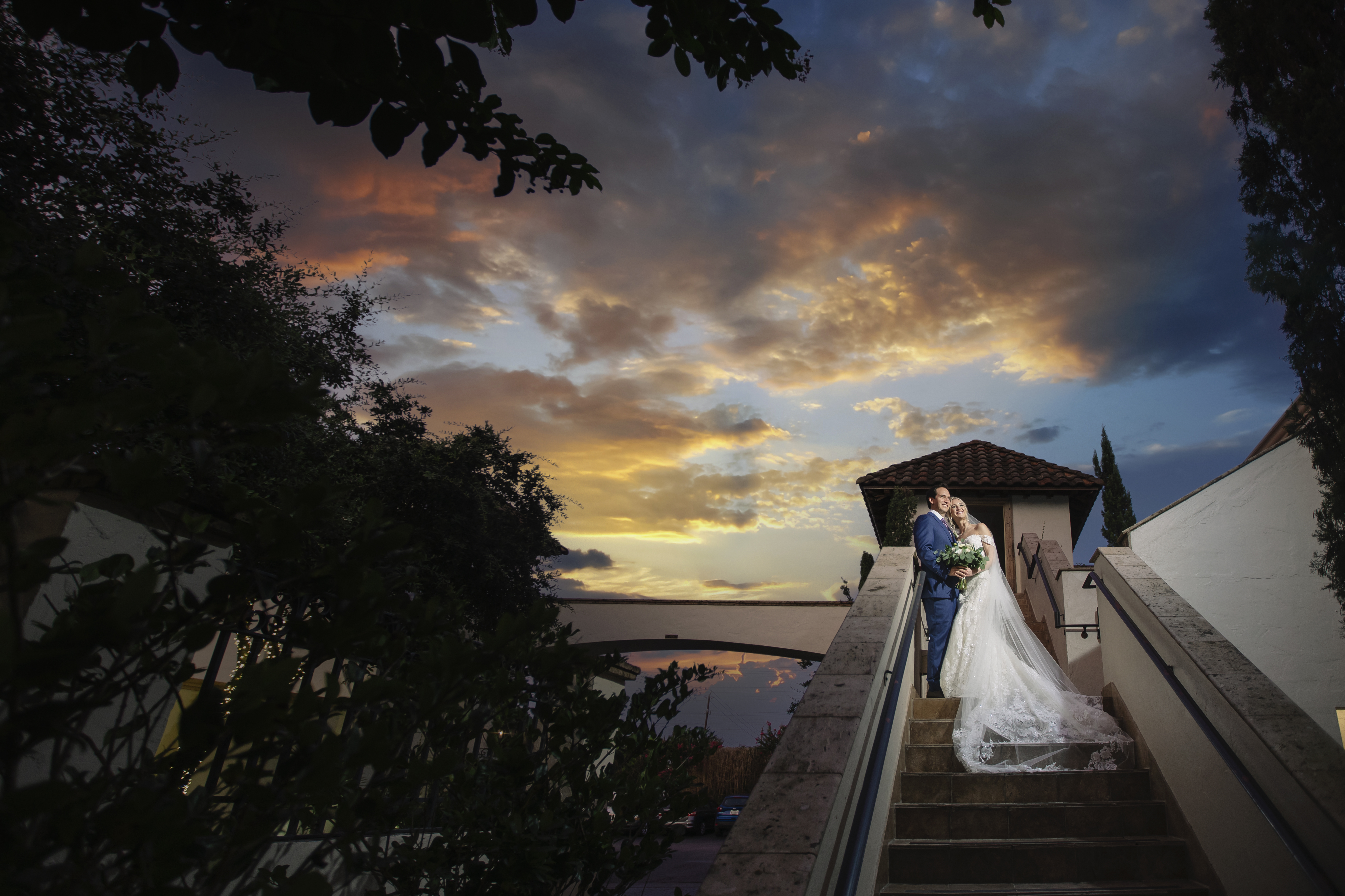 The bride and groom look up at the sky, which has a colorful sunset.