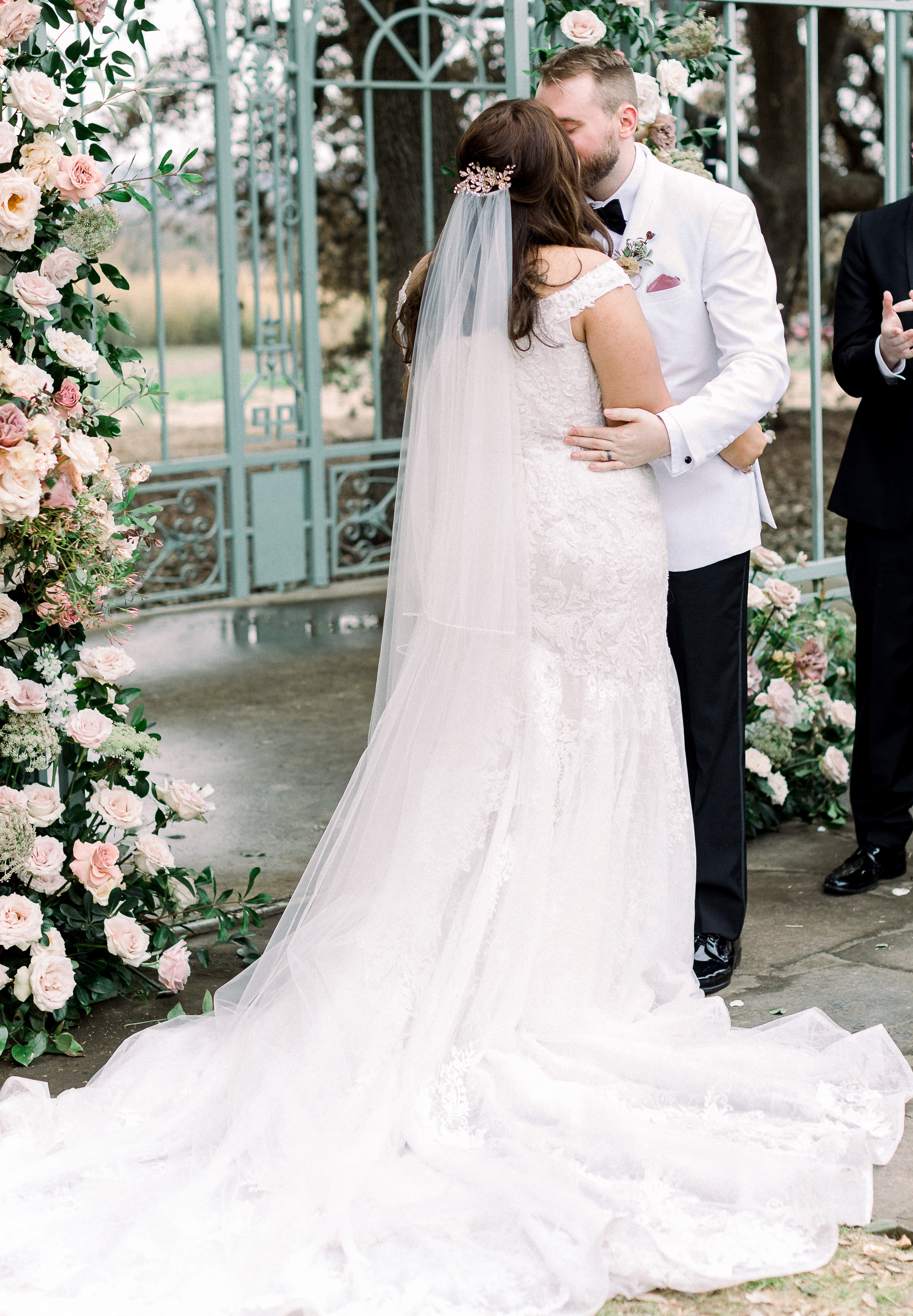 Bride, wearing long white veil and off the shoulder gown, and groom, in white tuxedo jacket and black pants,  kissing after an outdoor wedding ceremony at a hill country wedding venue. 