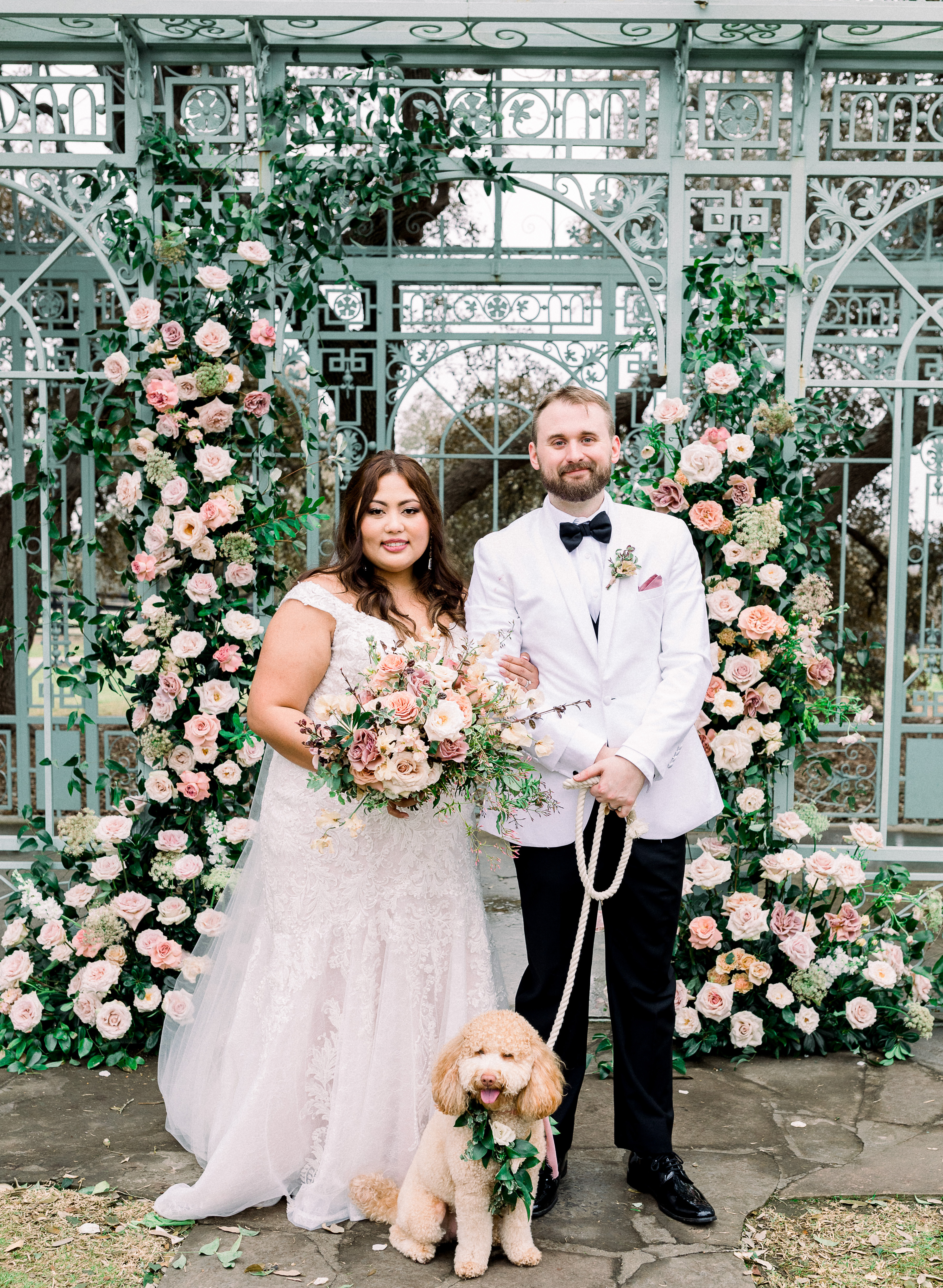The bride and groom stand in front of an antique french gazebo decorated with blush florals and greenery, with their pet miniature Goldendoodle, for their romantic al fresco wedding ceremony in Dripping Springs, Texas at wedding venue, Ma Maison.