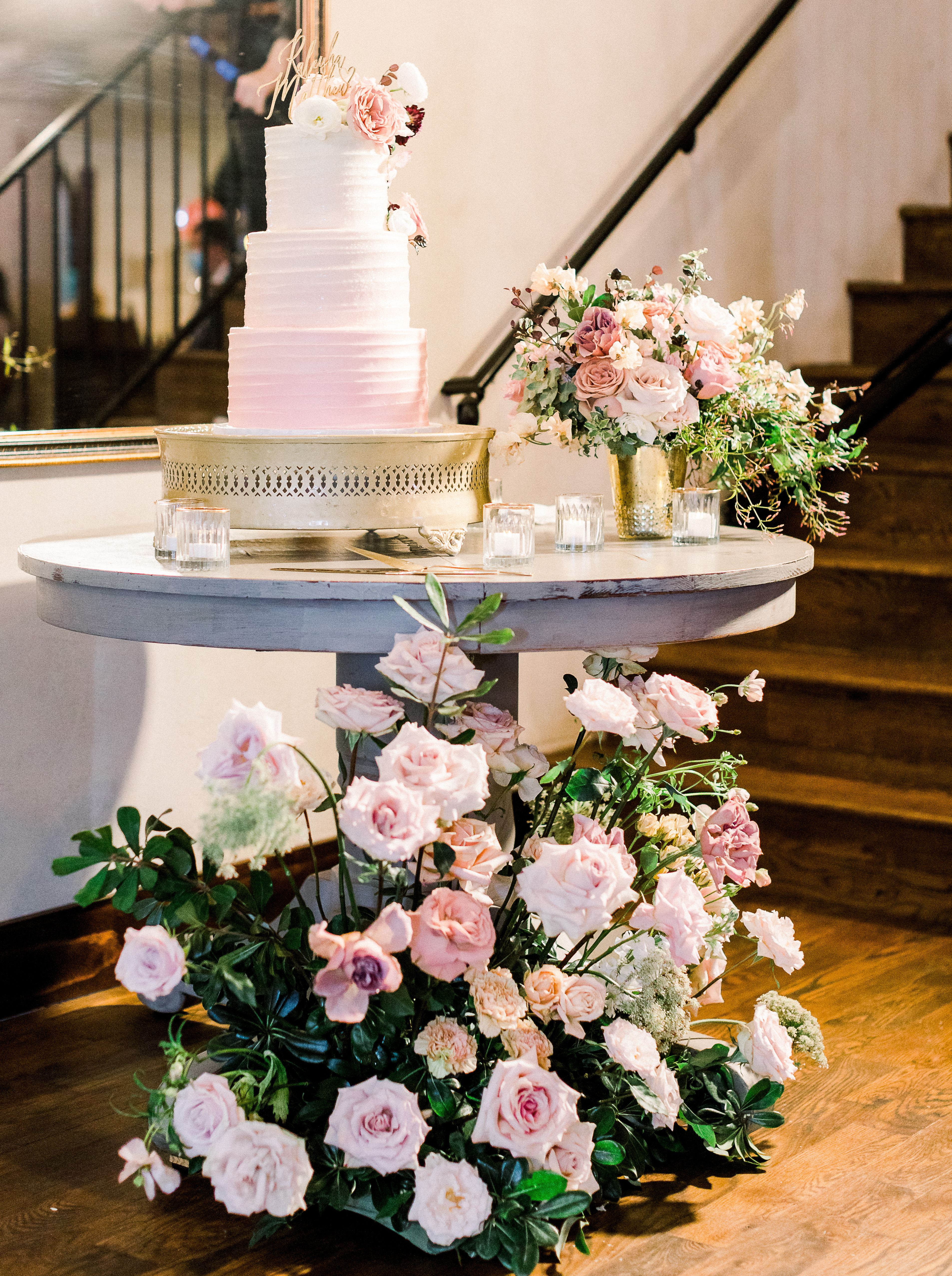 Pink ombre three tier cake by Sweet Treets Bakery atop a round table surrounded by fresh pink roses at a Texas hill country wedding reception. 