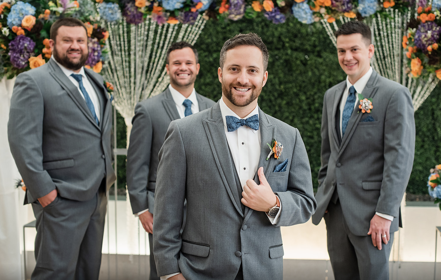 The groom smiles in front of the altar with a few of his groomsmen behind him.