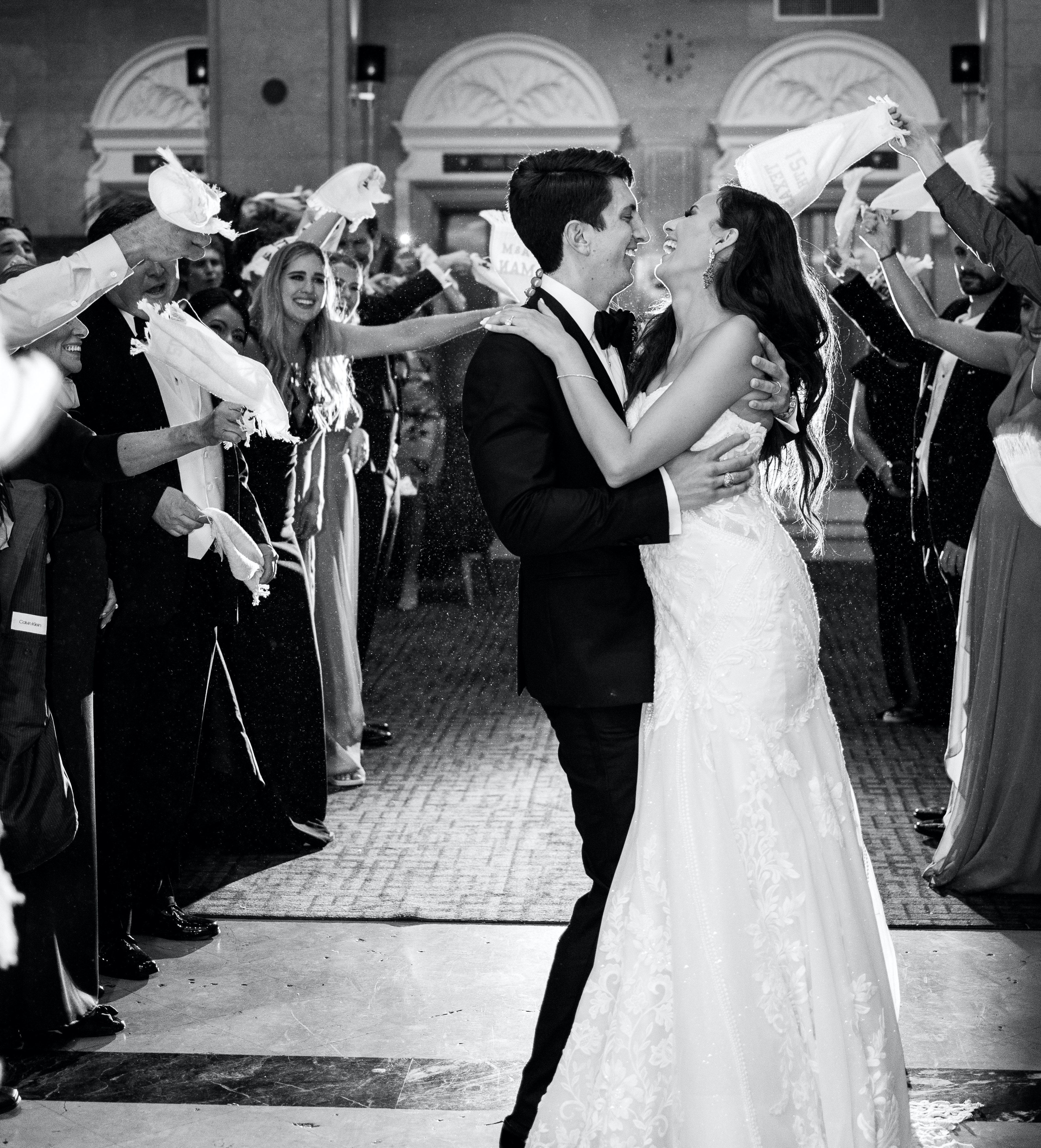 The bride and groom smile and hug each other while their guests wave towels in the air.