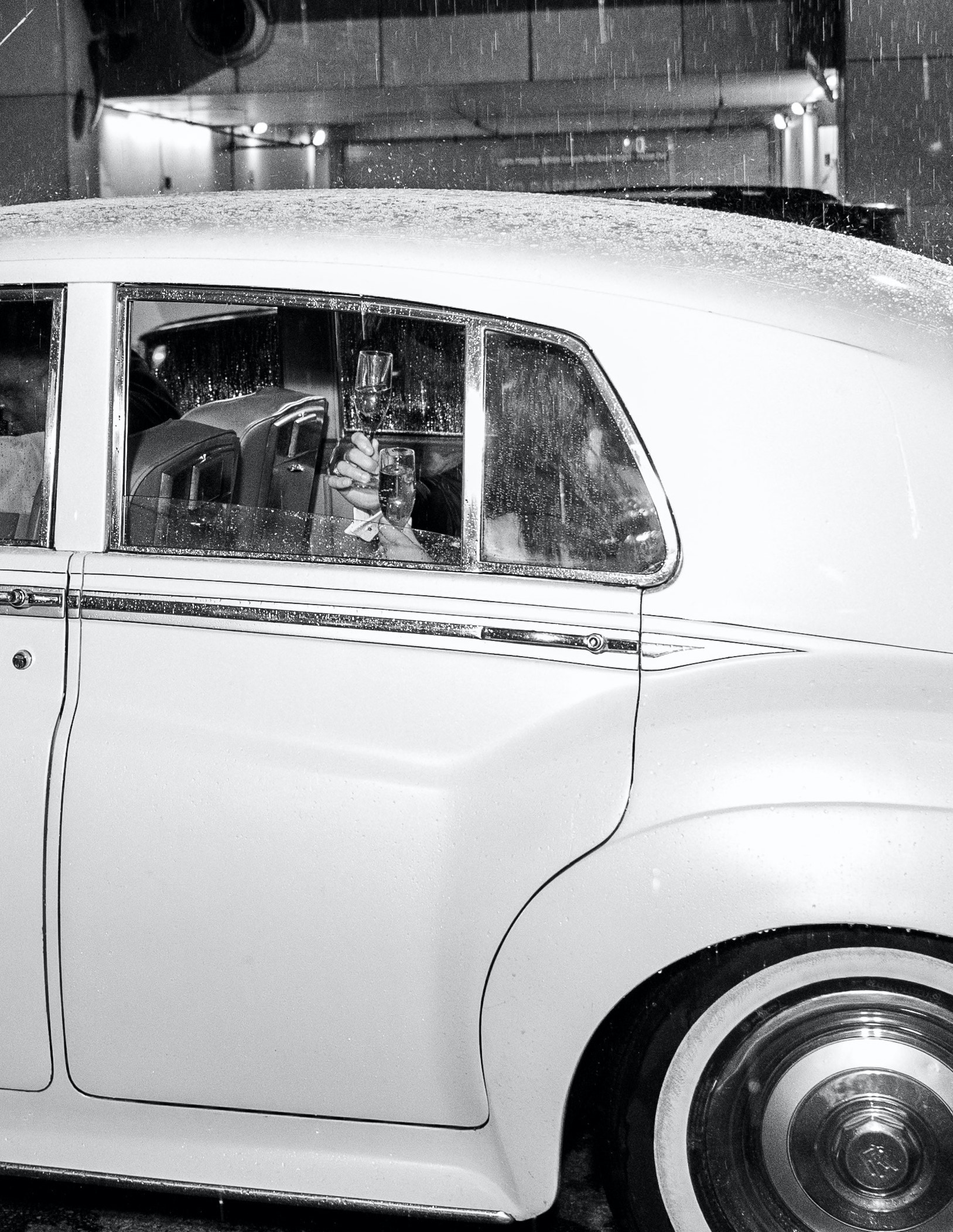 The bride and groom are sitting in the back of a Rolls Royce clinking champagne glasses.