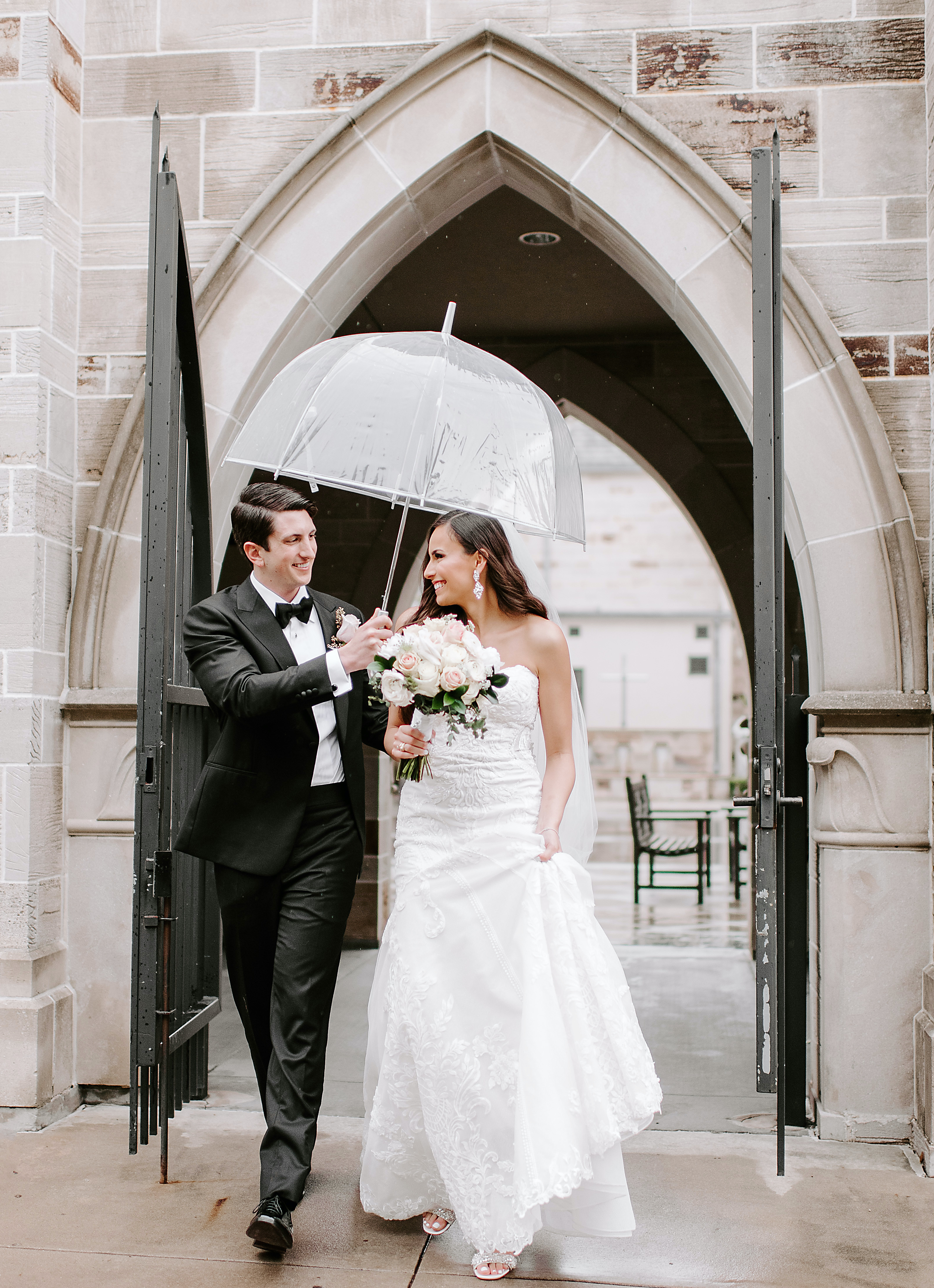 The groom holds a clear umbrella over the bride's head after their elegant rainy wedding.