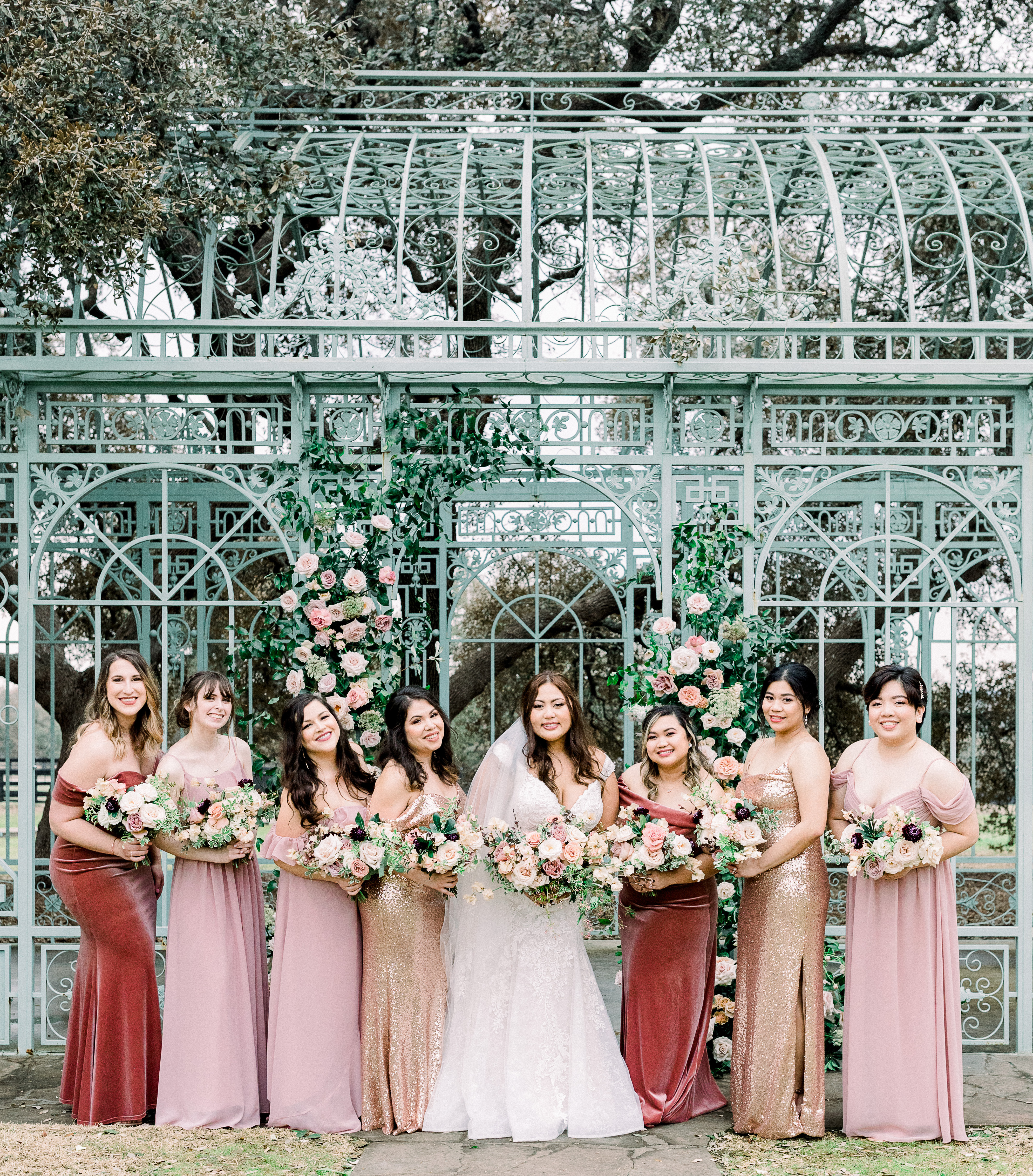Bride holding bouquet surrounded seven bridesmaids in monochromatic pink, rust and gold dresses in front of an antique french gazebo at hill country wedding venue, Ma Maison.