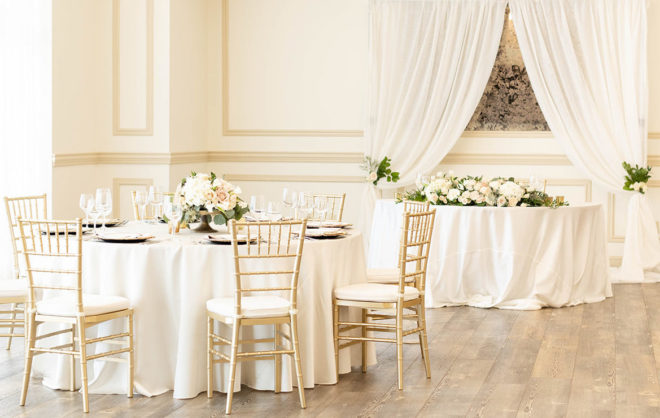 Indoor reception hall, Kendall Halle, with round table with gold chivari chairs and a sweetheart table with white linens set with florals at The Kendall in Boerne, Texas. 