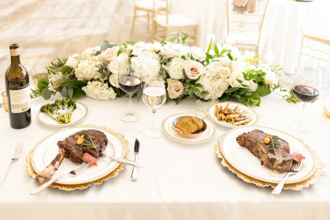 Two place settings with beef tenderloin, grilled broccoli, carrots and wine glasses filled with red wine on a small sweetheart table set with white and pink florals in the Kendall Halle, a wedding event space at The Kendall Hill Country Inn in Boerne, Texas. 