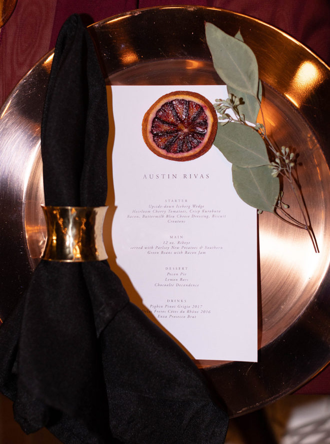 Gold plate set with black napkin, an orange rind, and grennery plus a menu that reads, "Austin Rivas" "Starters" "Main" "Dessert" and "Drinks" with menu items from Boerne restaurant, Peggys on the Green.