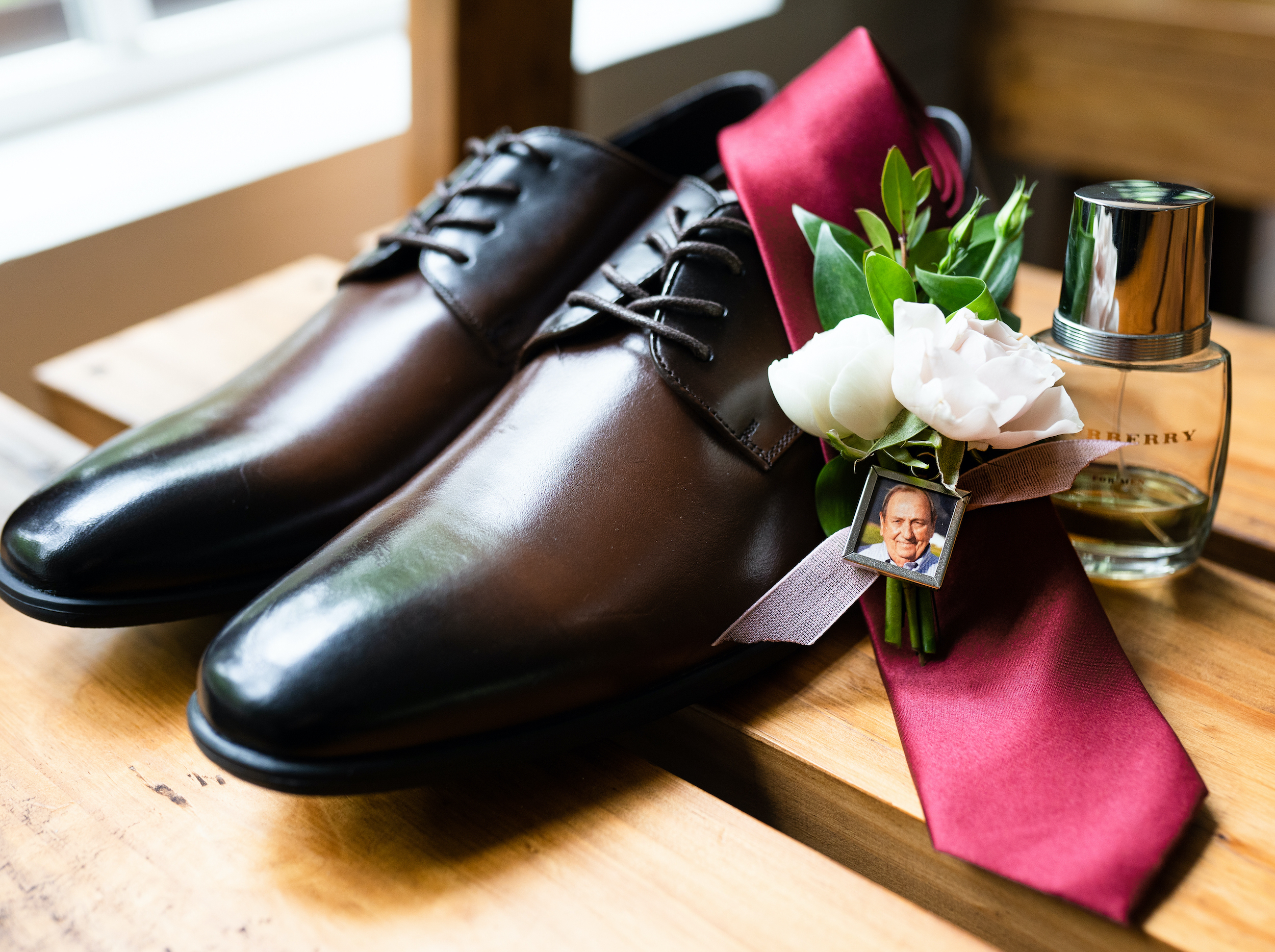 The groom's shoes and burgundy tie are placed next to his cologne and a photo of a family member.