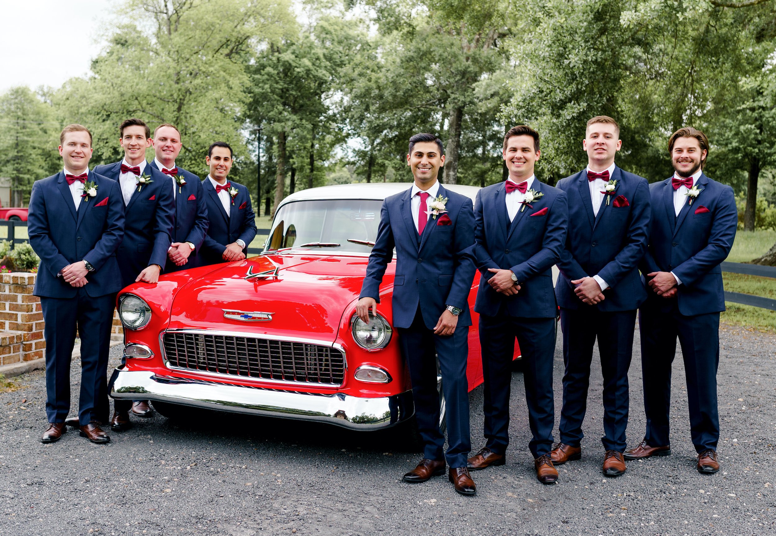 The groom and groomsmen smile in their navy tuxes next to the bright red classic car.