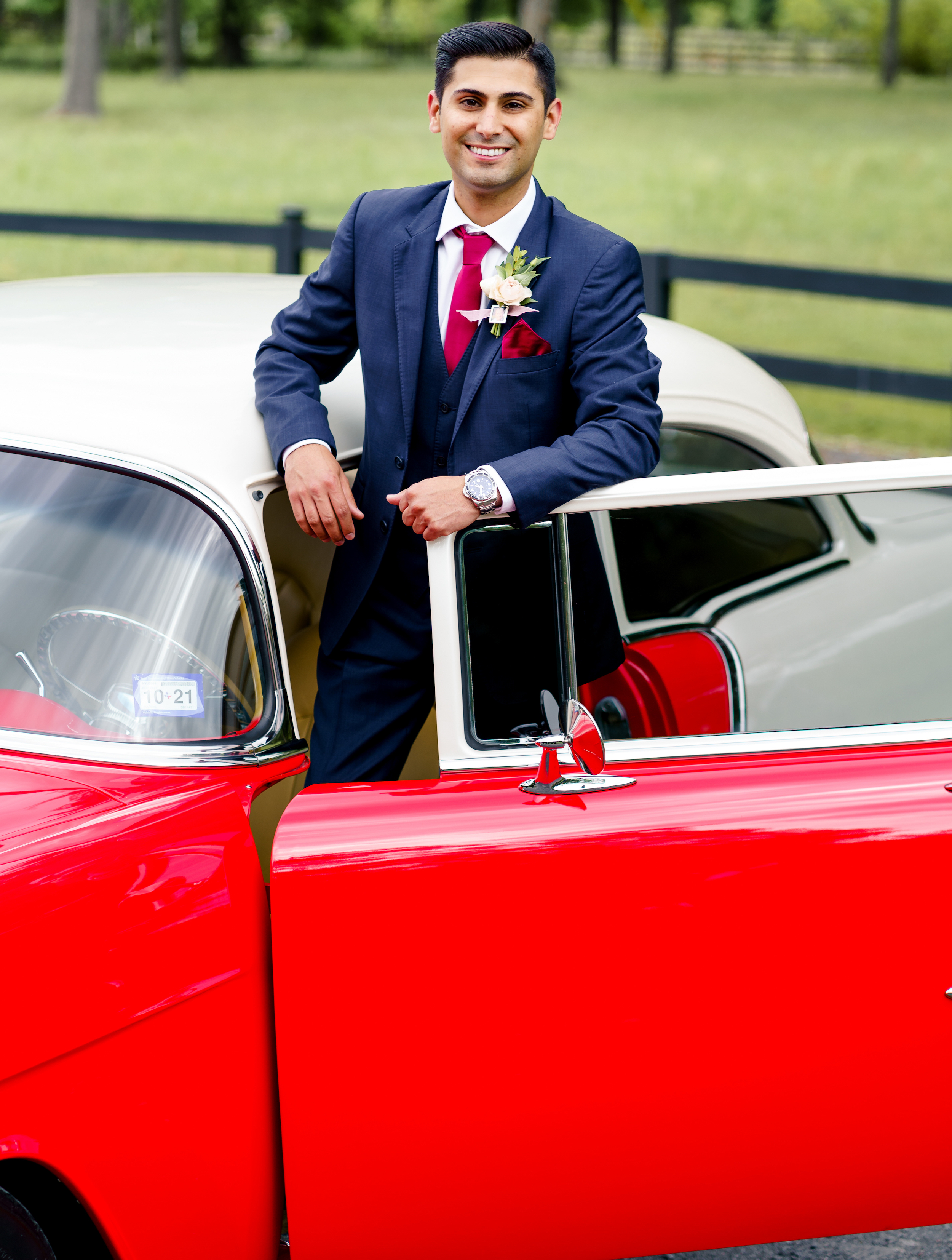 The groom is standing outside of the driver's seat of a bright red classic car parked on The Carriage House property in Montgomery, TX.