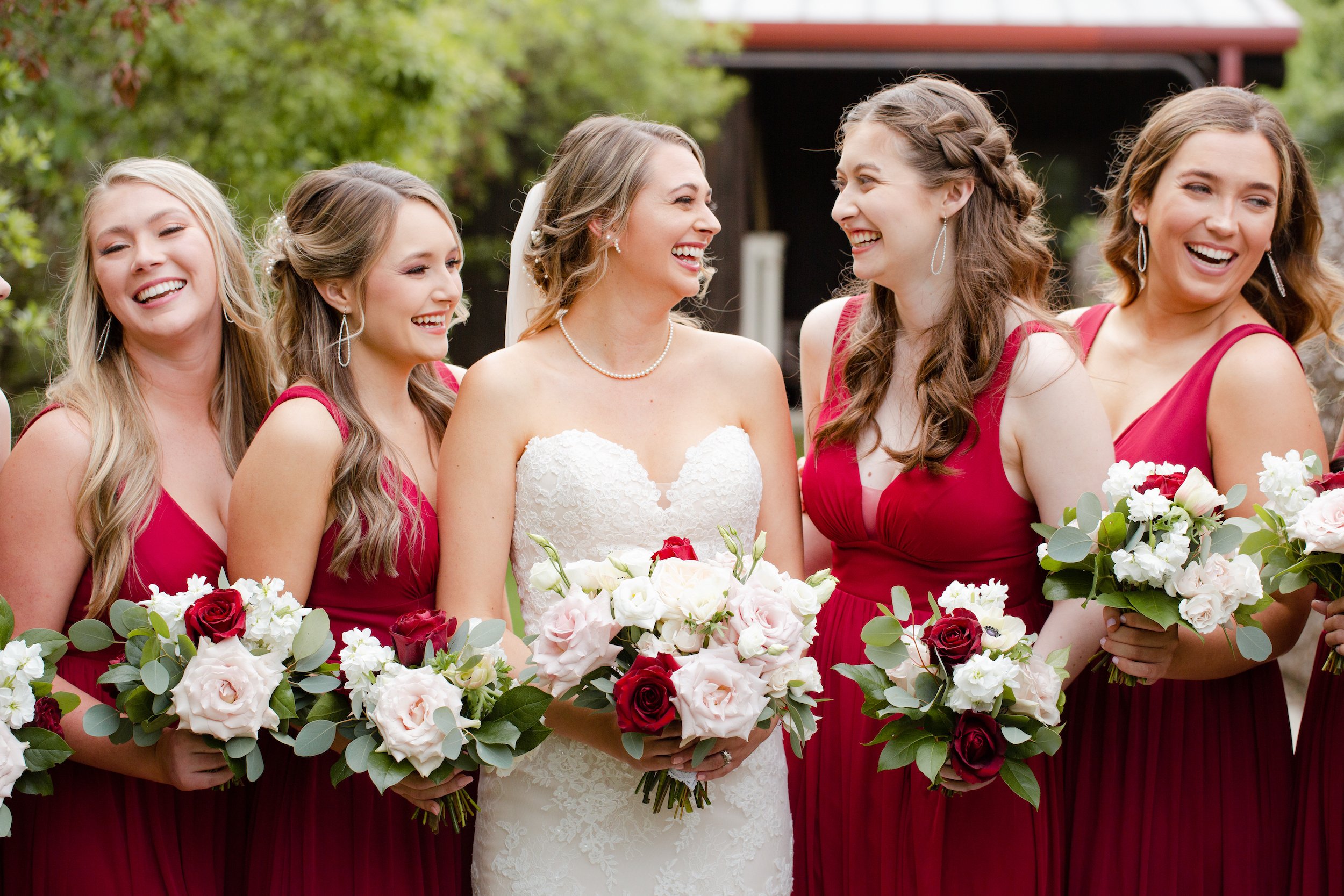 The bride smiles with her bridesmaids and they are all holding their burgundy and blush flower bouquets.