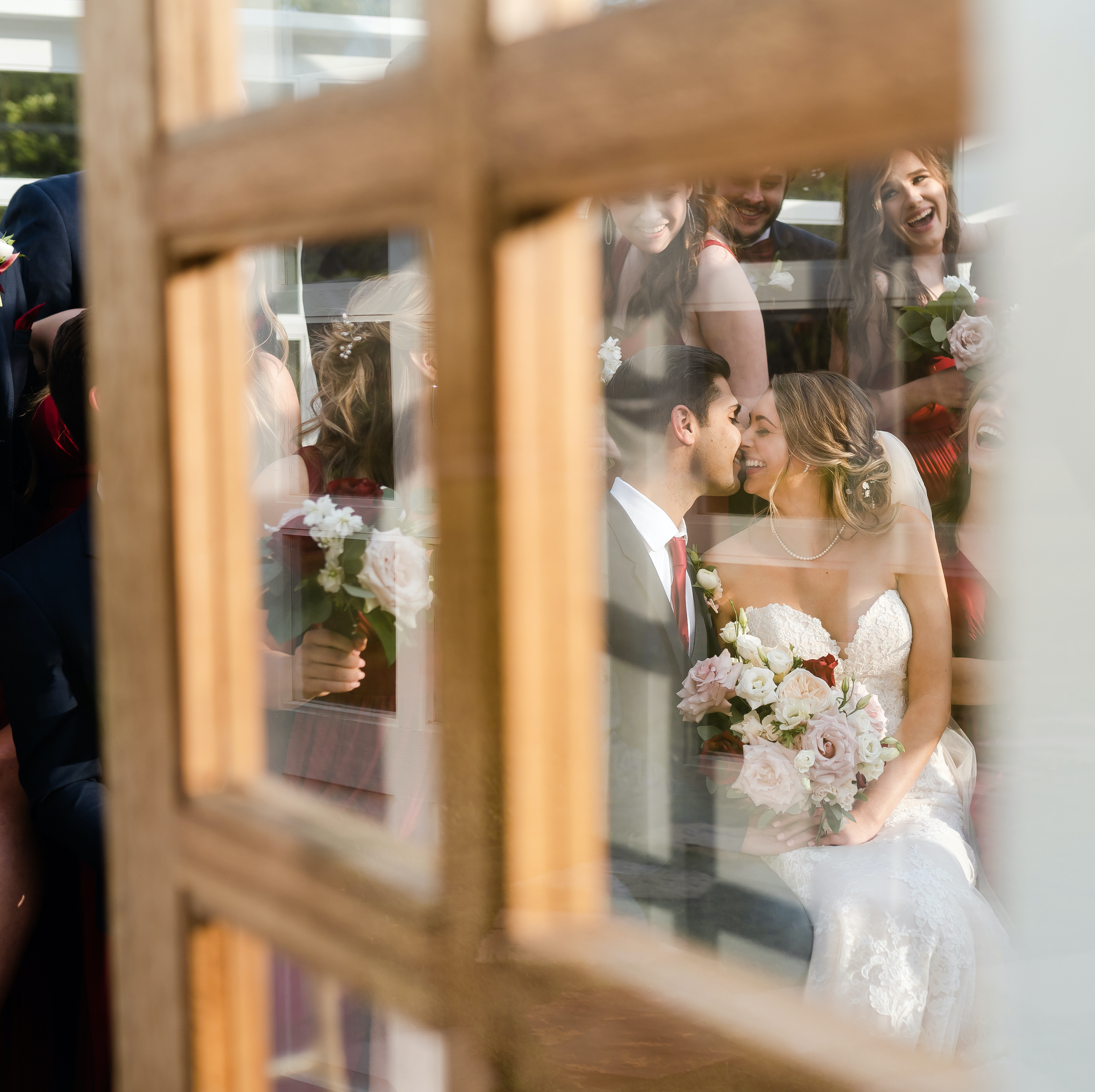 Through the window of a door, you can see the bride and groom smile and kiss while sitting with their wedding party.