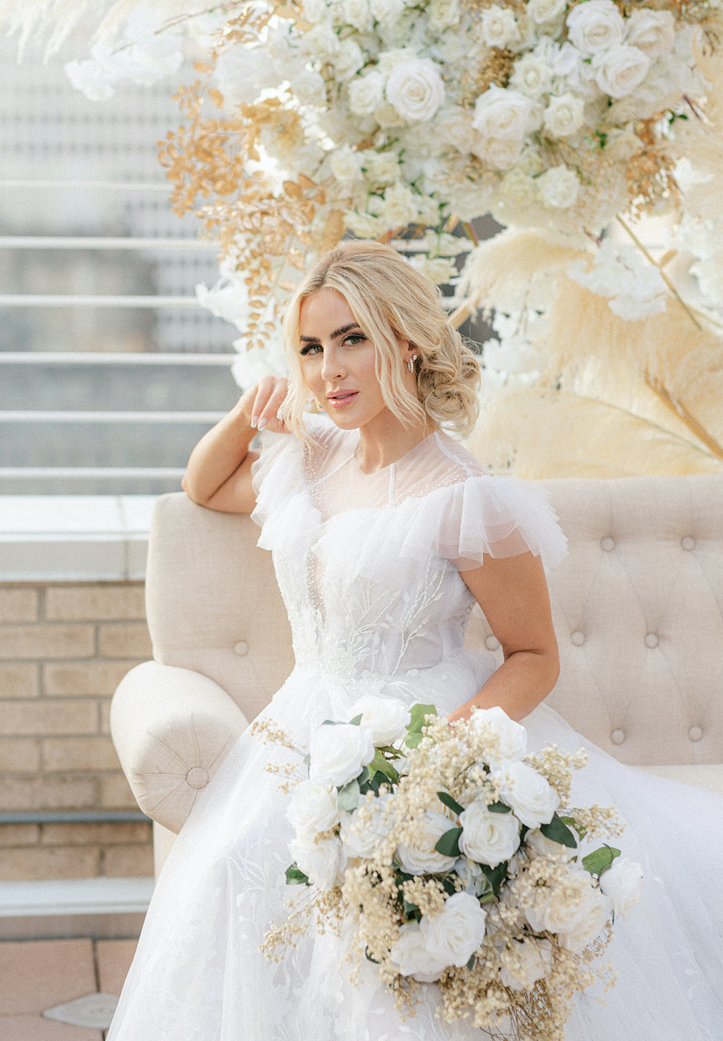 The bride poses on the beige couch, holding her white rose bridal bouquet for the ethereal rooftop wedding styled shoot.