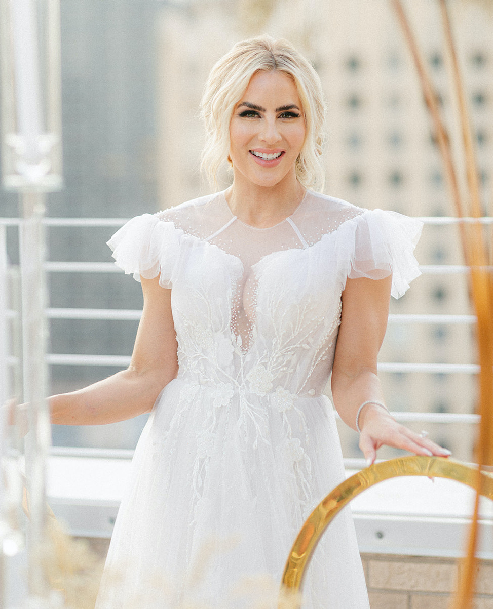 The bride stands between the gold chairs at the dinner table, smiling at the camera in her wedding gown.