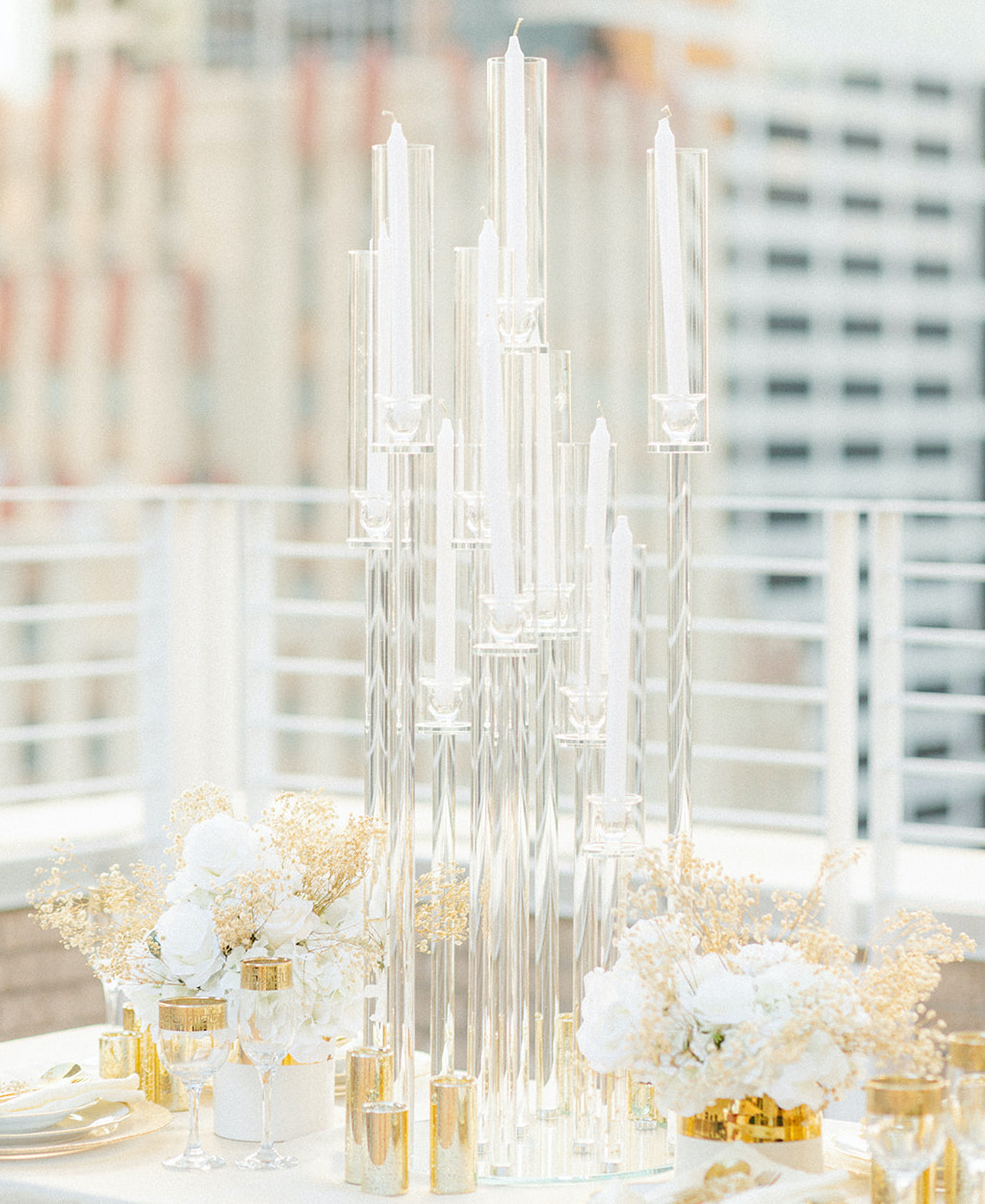 A close up photo of the tall candle votives that are translucent glass. Gold accented flower arrangements and dinnerware surround the tall candles.