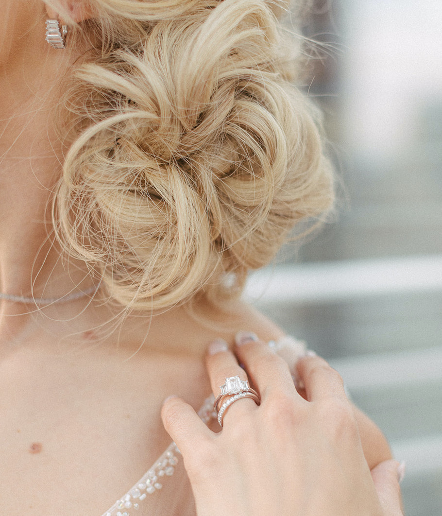 The bride is wearing a low lose bun and has her hand on her shoulder with her engagement ring on.